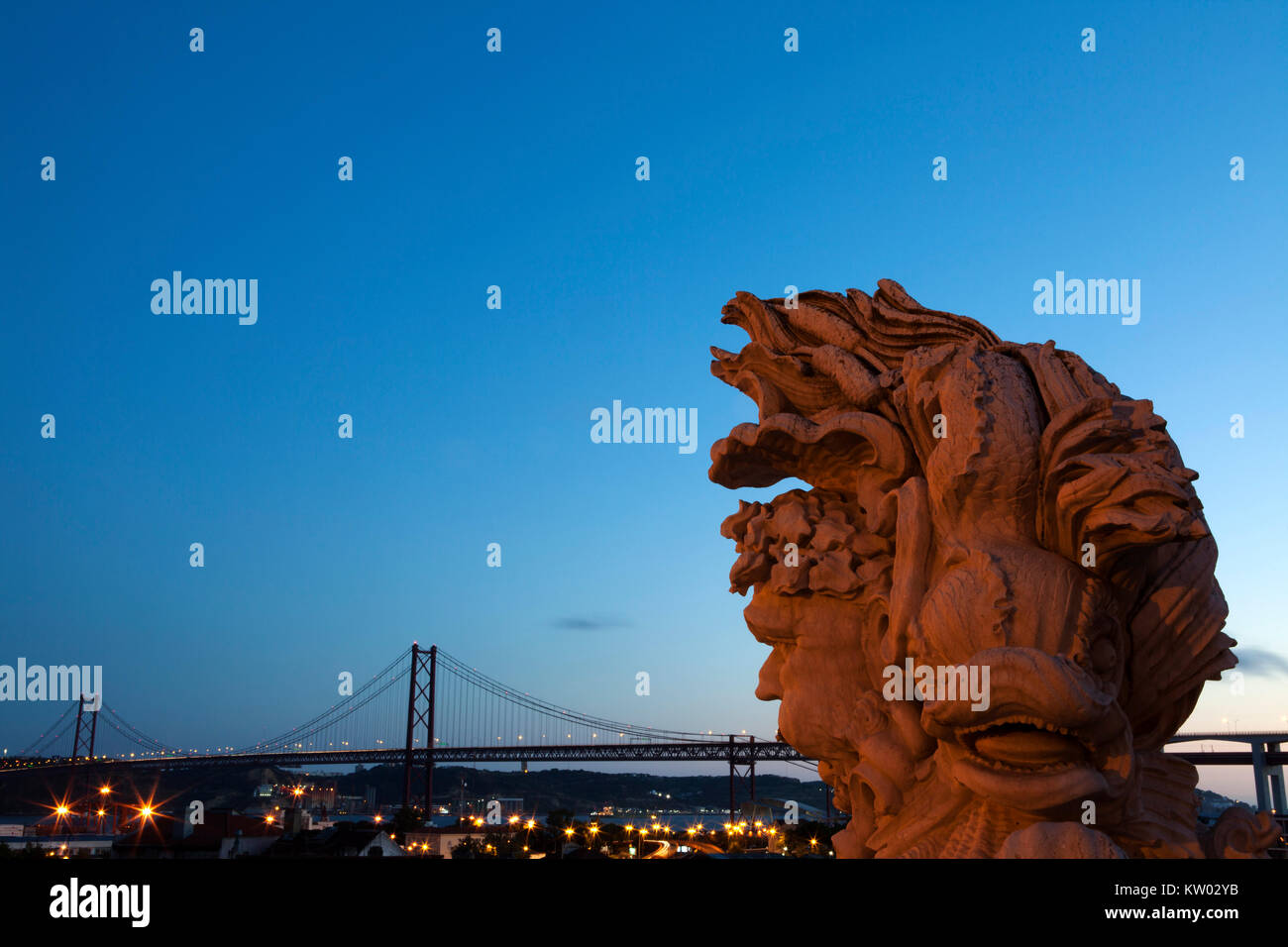 View of the 25 April Bridge (Ponte 25 de Abril) in Lisbon, Portugal. A sculpture stands in the foregrounds at the Miradouro do Largo das Necessidades. Stock Photo