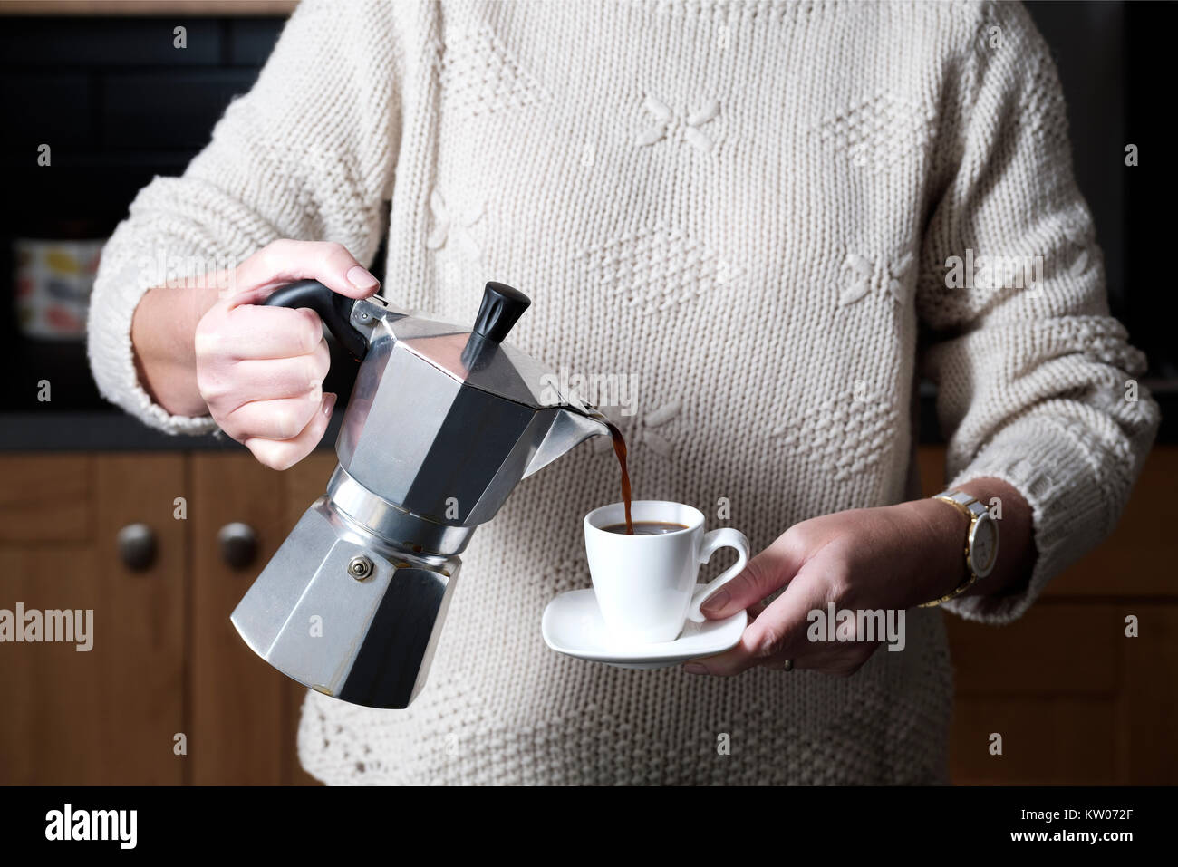 A smart but casually dressed woman pouring an espresso coffee from a moka hob espresso maker whilst standing in her kitchen at home Stock Photo