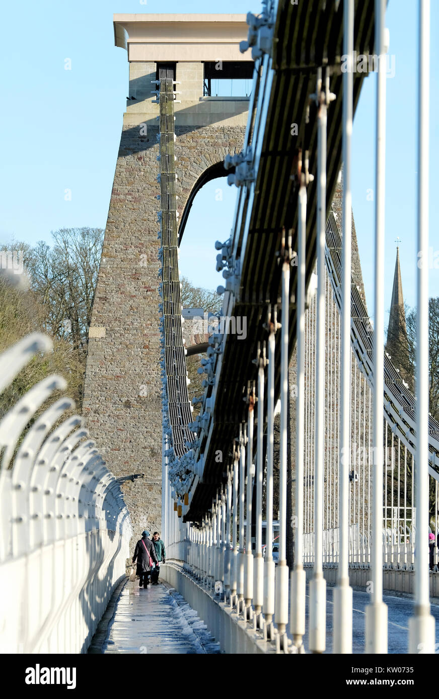 A woman carrying a large bag walking across the Clifton Suspension Bridge in Bristol, UK. A cold winter day and there is ice and snow on the footpath. Stock Photo