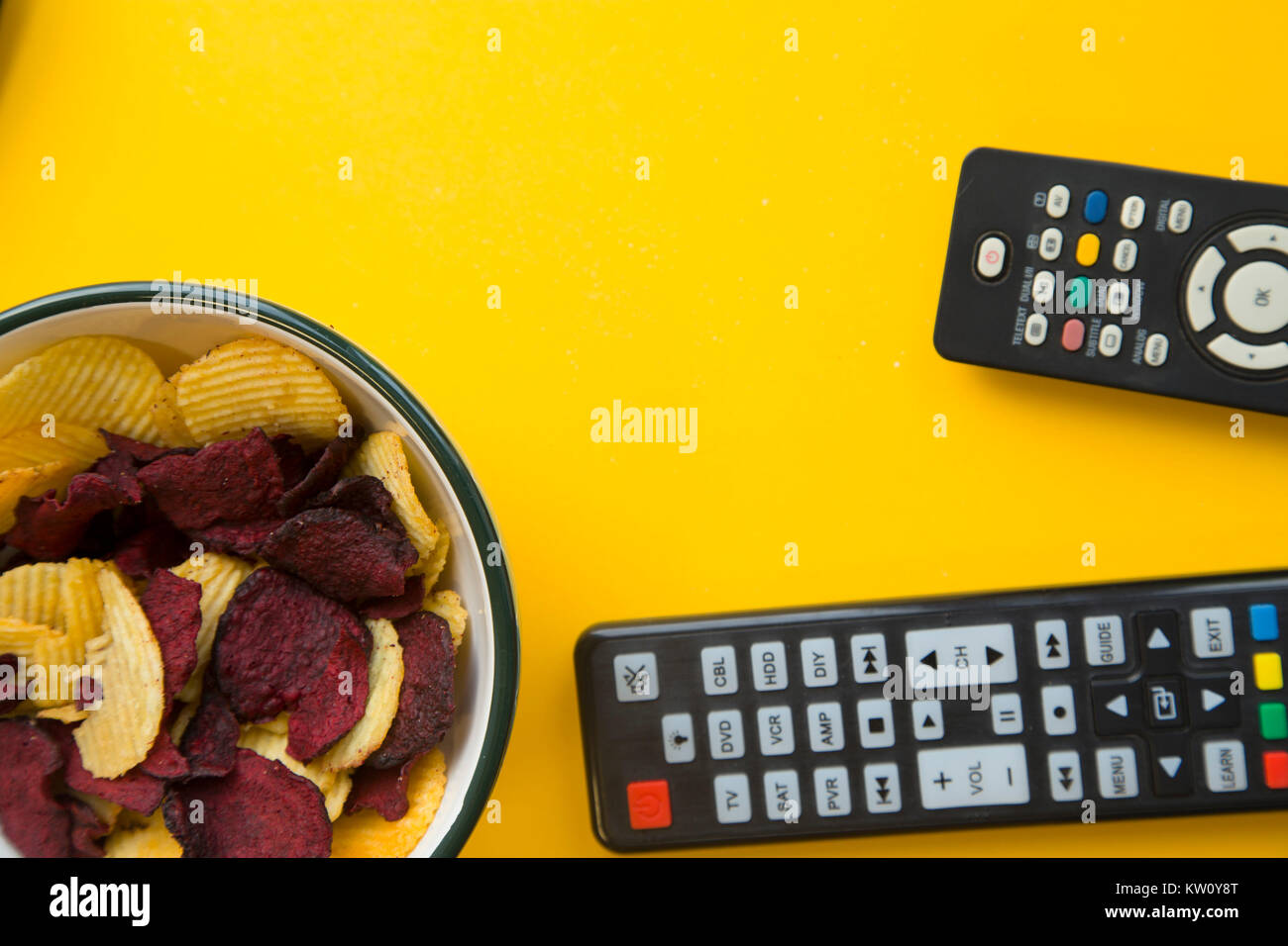 Relaxing in front of the tv. Weekend, hobby and leisure concept. A bowl of potato and beetroot chips and two remote controls on a bright yellow backgr Stock Photo