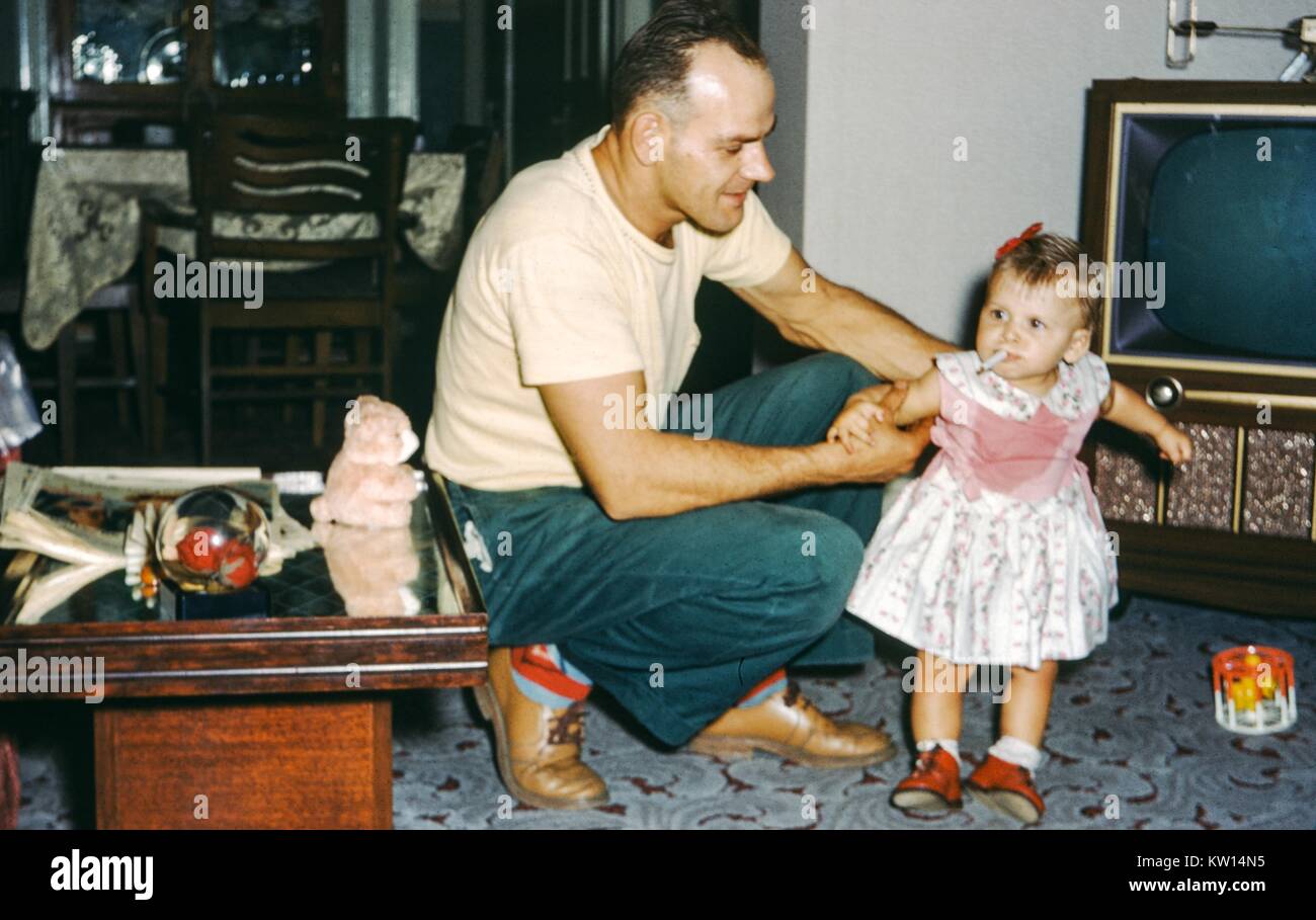 Father kneeling down in a suburban living room and holding his young  daughter, who has a cigarette in her mouth and is pretending to smoke, 1952  Stock Photo