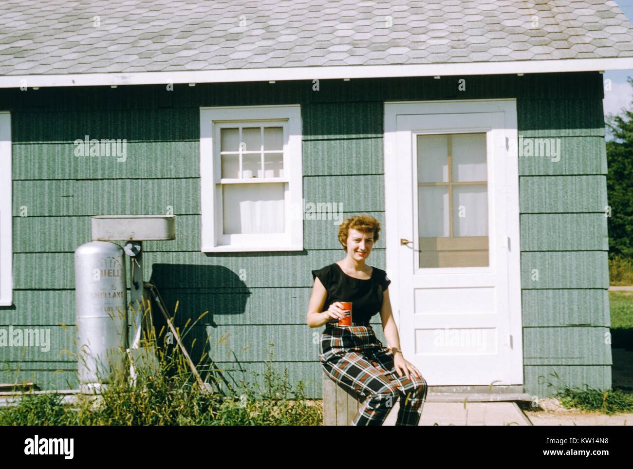 Red-haired woman sitting outside a mountain camping cabin and holding a can of soda, with Shell propane tank visible in the background, 1952. Stock Photo