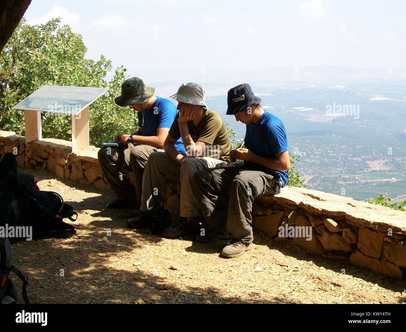 On Mount Benthal, three Israel hikers wearing matching khaki military pants and wide-brimmed hats rest on a stone wall, with their backpacks visible in the foreground, Mount Benthal, Golan Heights, Israel, 2012. Stock Photo