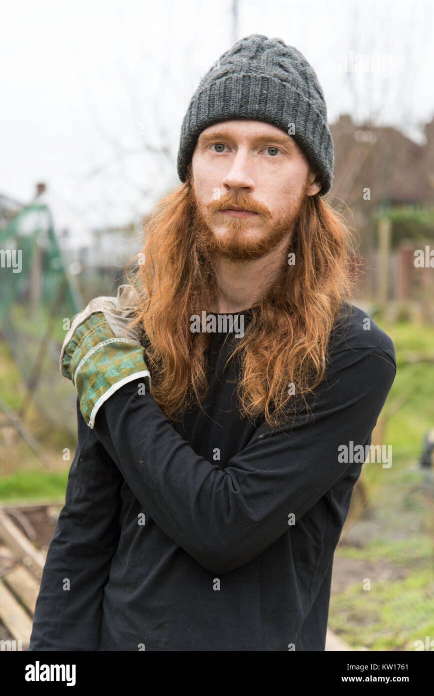 A portrait of a male gardener posing in an allotment with long ginger hair, beard and moustache wooly hat, black top and gardening gloves. Stock Photo