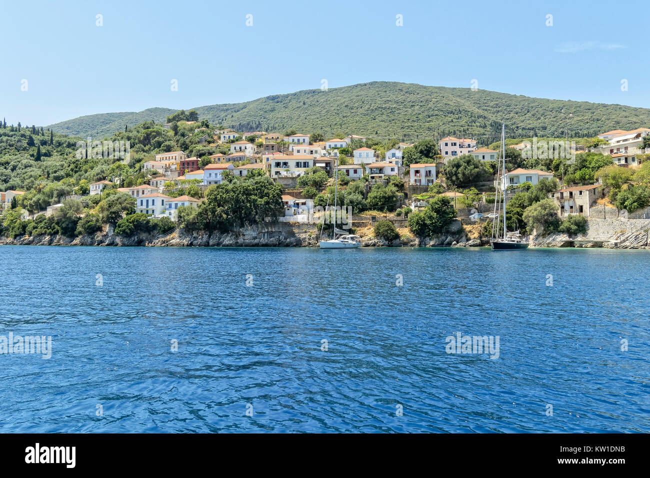 On the shores of the Greek island of Ithika white painted houses nestle among trees on the hillside while two yachts are moored close to the shore. Stock Photo