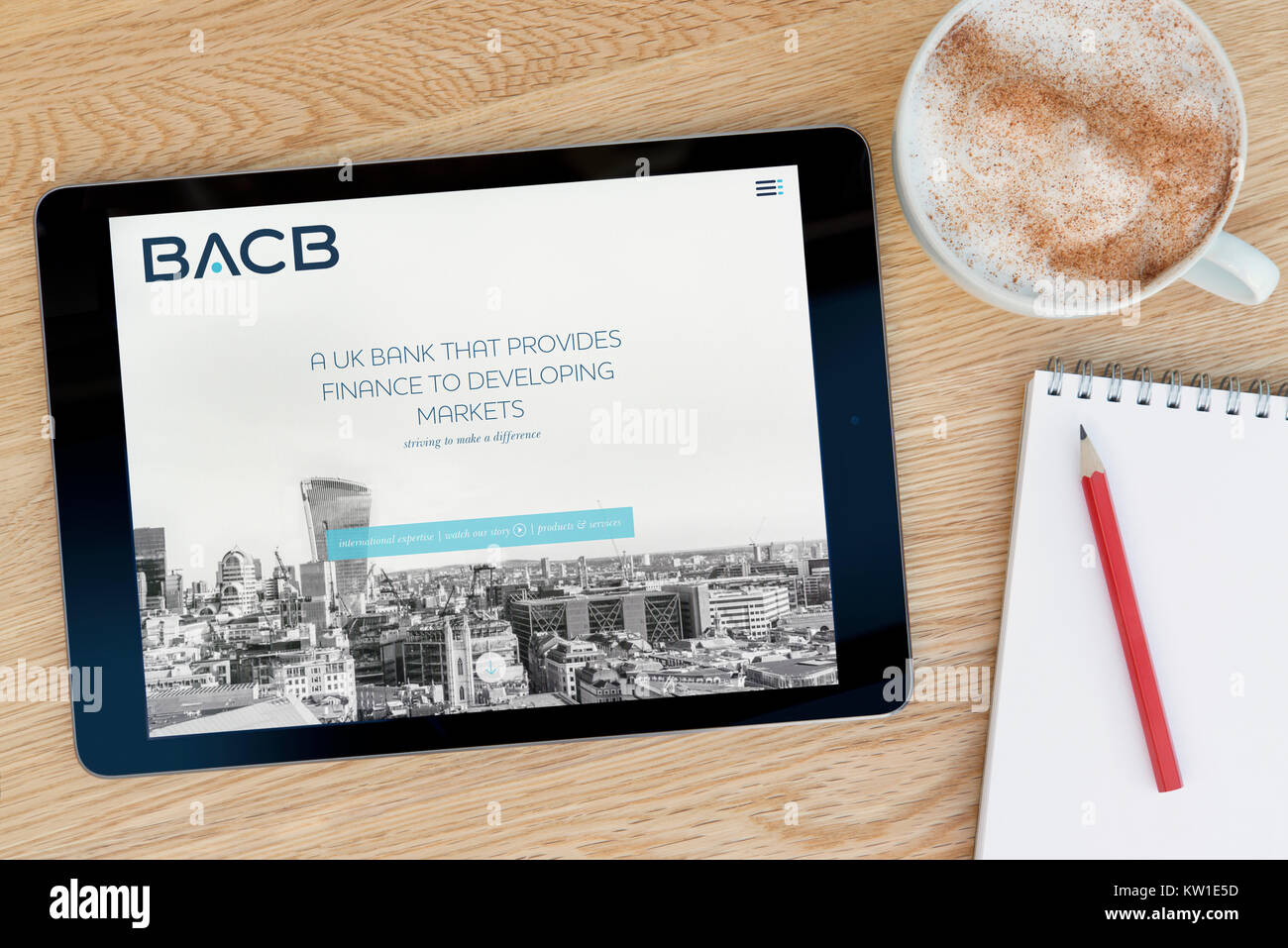 The BACB (British Arab Commercial Bank) website on an iPad, resting on a wooden table beside a notepad, pencil and cup of coffee (Editorial use only). Stock Photo