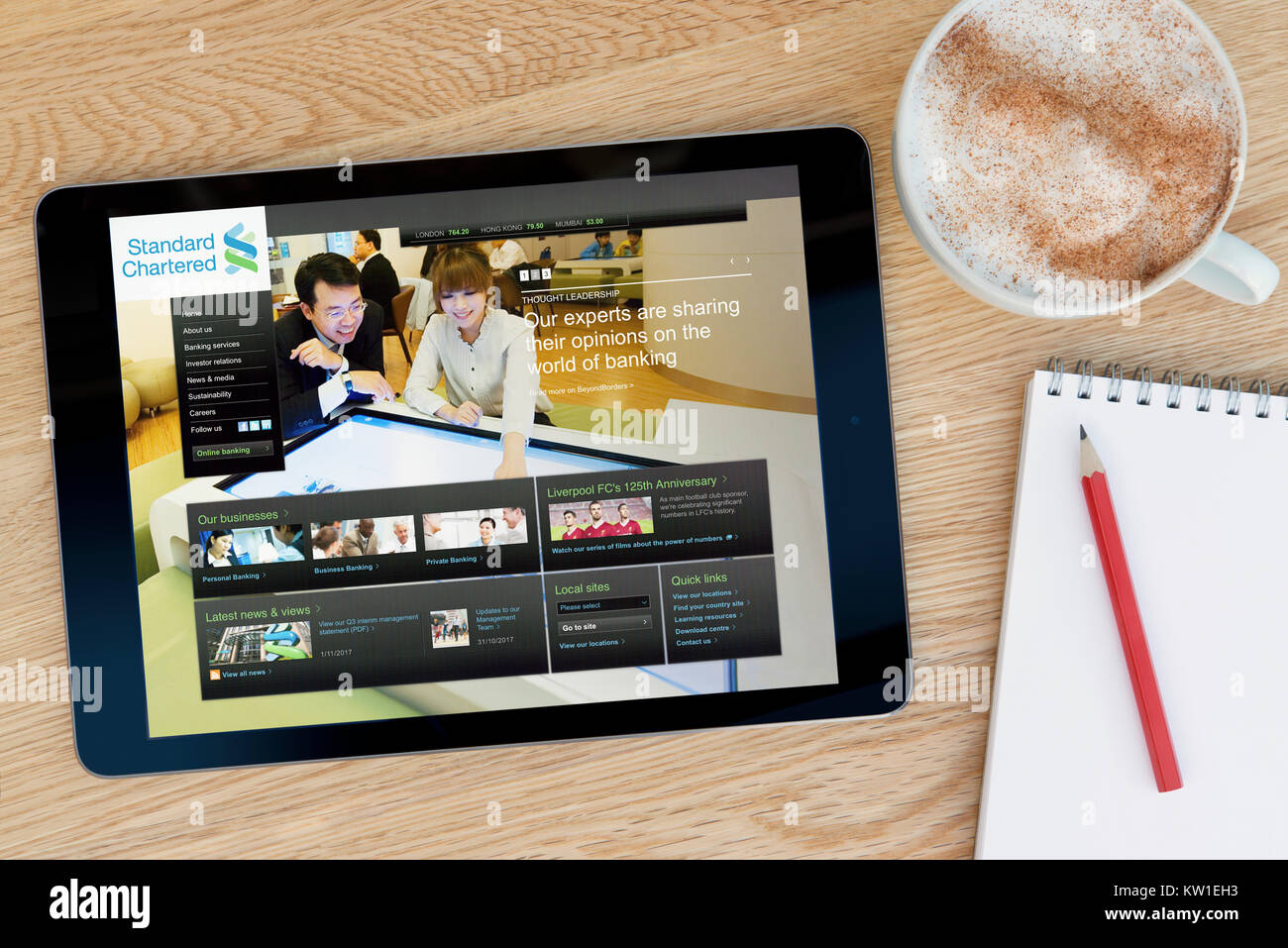 The Standard Chartered website on an iPad tablet device, resting on a wooden table beside a notepad, pencil and cup of coffee (Editorial only) Stock Photo