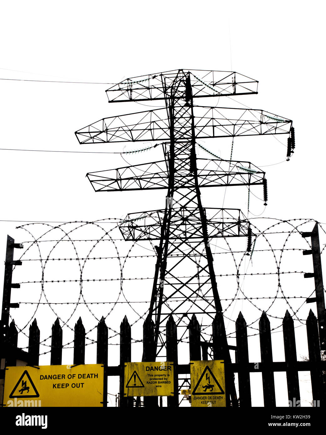 Silhouette of an electricity pylon against a grey sky with razor wired security fence in foreground Stock Photo