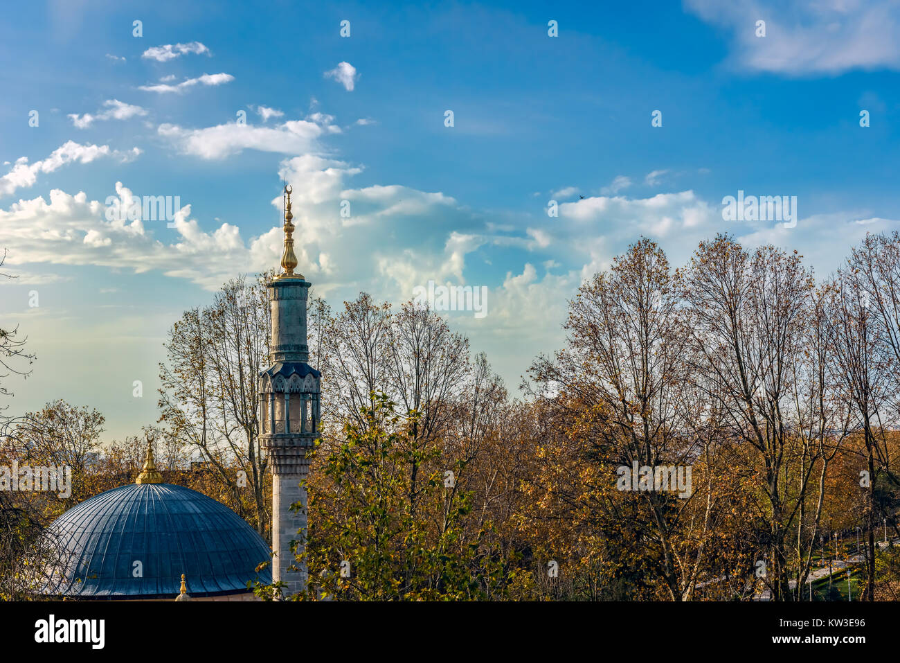 A mosque dome and its minaret in front of a grove with fallen reddish leaves under beautiful blue sky with white fluffy clouds Stock Photo