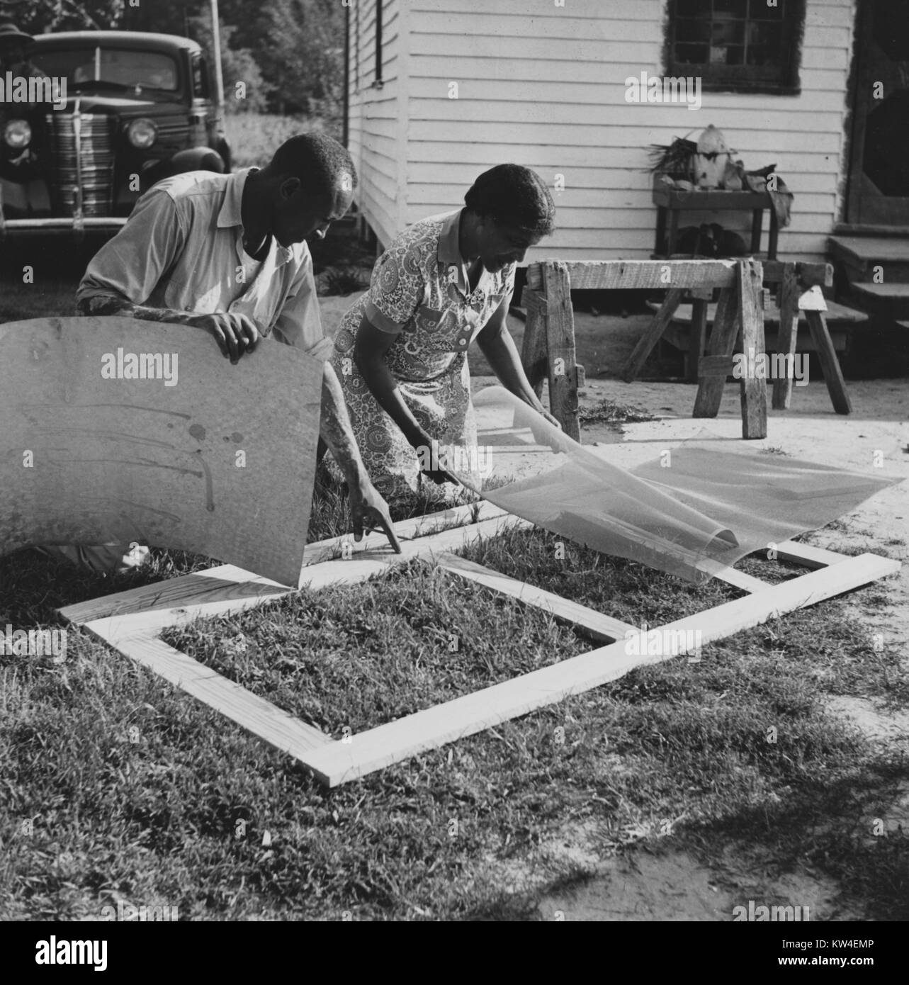 A man and woman kneel in the grass in front of a house demonstrating home screen door construction in Ridge, Saint Mary's County, Maryland, July, 1941. Stock Photo