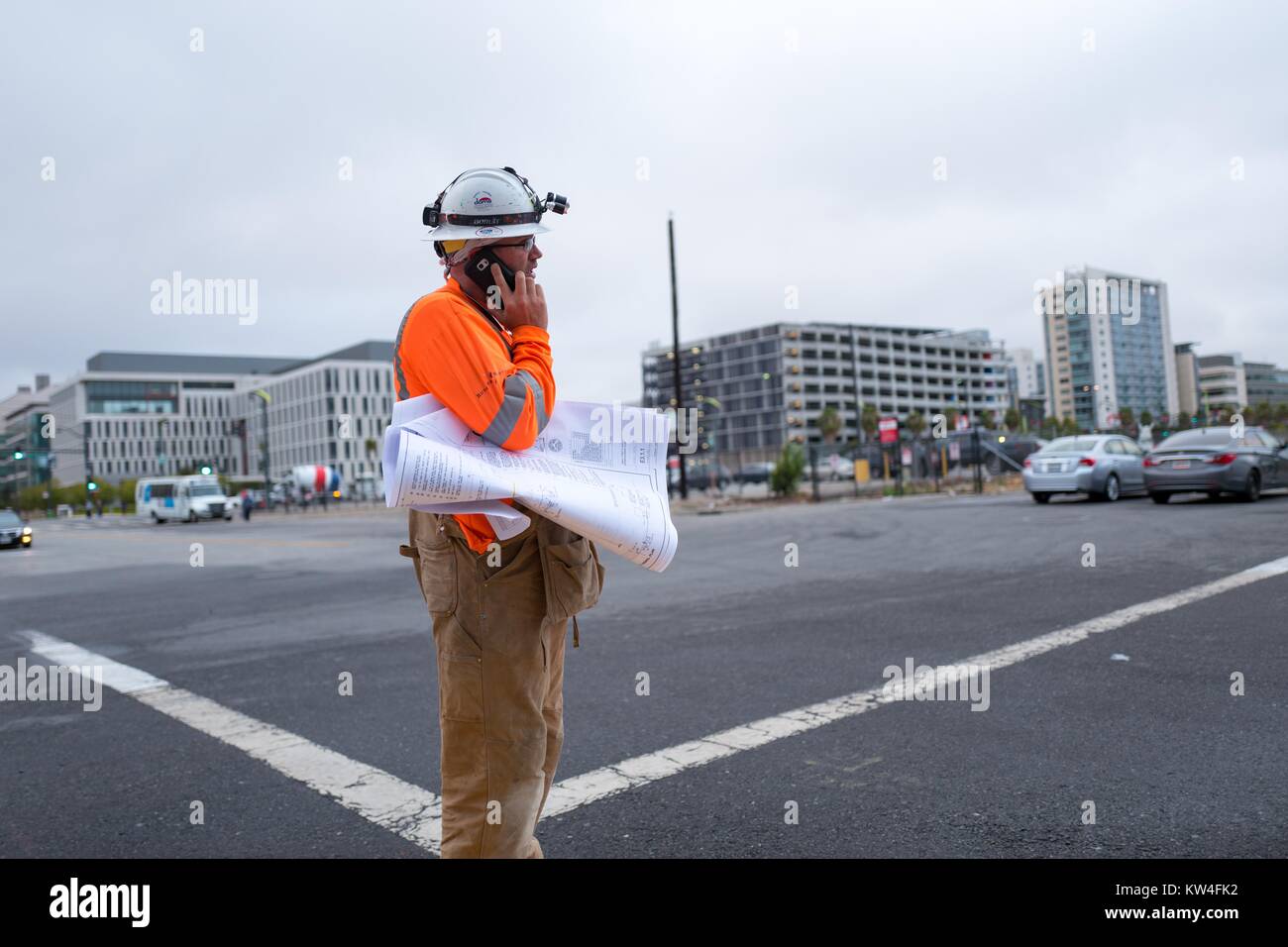 In the Mission Bay neighborhood of San Francisco, California, a construction foreman stands on a street corner and speaks on a cellphone, while holding blueprints under his arm and wearing a high-visibility vest and hard hat, San Francisco, California, August, 2016. Stock Photo