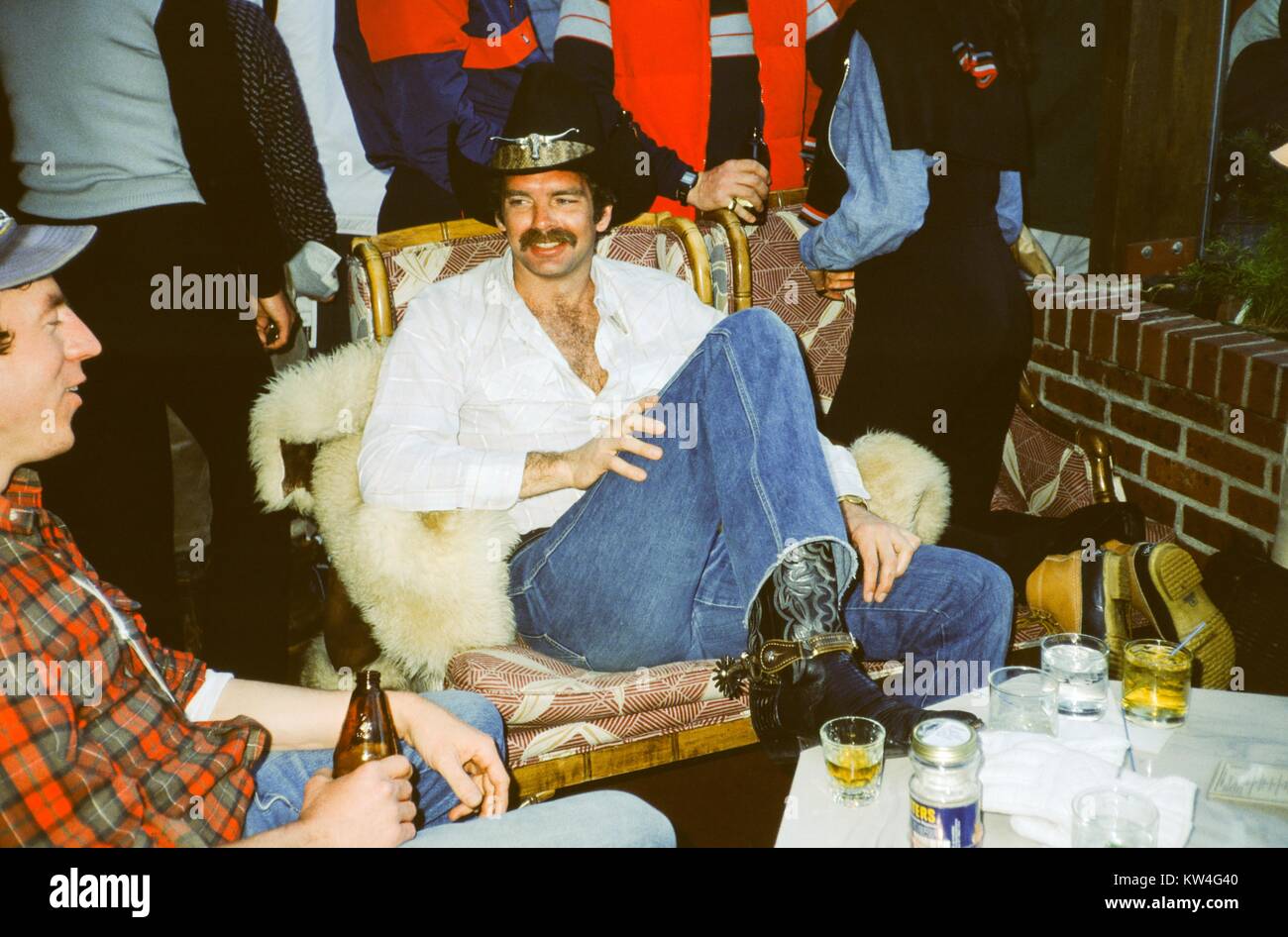 Man in cowboy hat and spurs, with a white shirt partially unbuttoned, sits with his feet up on a table, speaking and drinking alcohol with a man wearing a plaid shirt, 1975. Stock Photo