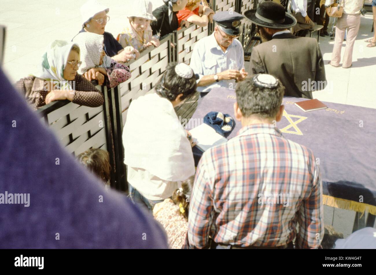 At the Western Wall (kotel) in Jerusalem, Israel, several women wearing hats and shawls peer over the divider between the male and female sections of the wall, as Jewish men wearing yarmuckles and tallit pray, 1975. Until 2016, the Western Wall was divided according to gender. Stock Photo