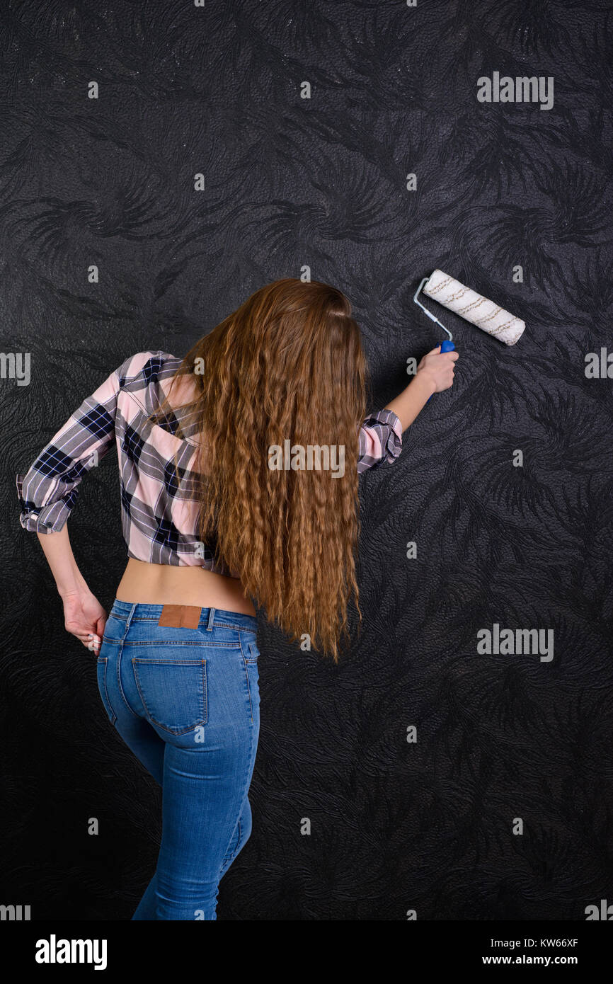 A girl in blue jeans and plaid shirt is holding paint roller near black textured wall Stock Photo