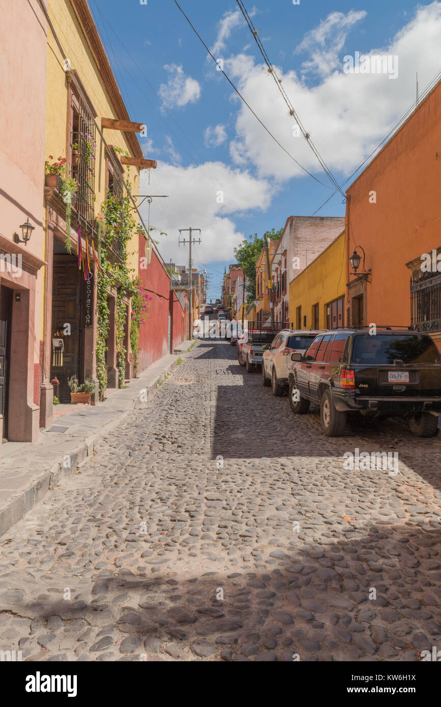 Colorful shops, and houses, on cobblestone street, with a cloudy blue sky and some parked cars, in San Miguel de Allende, Mexico Stock Photo