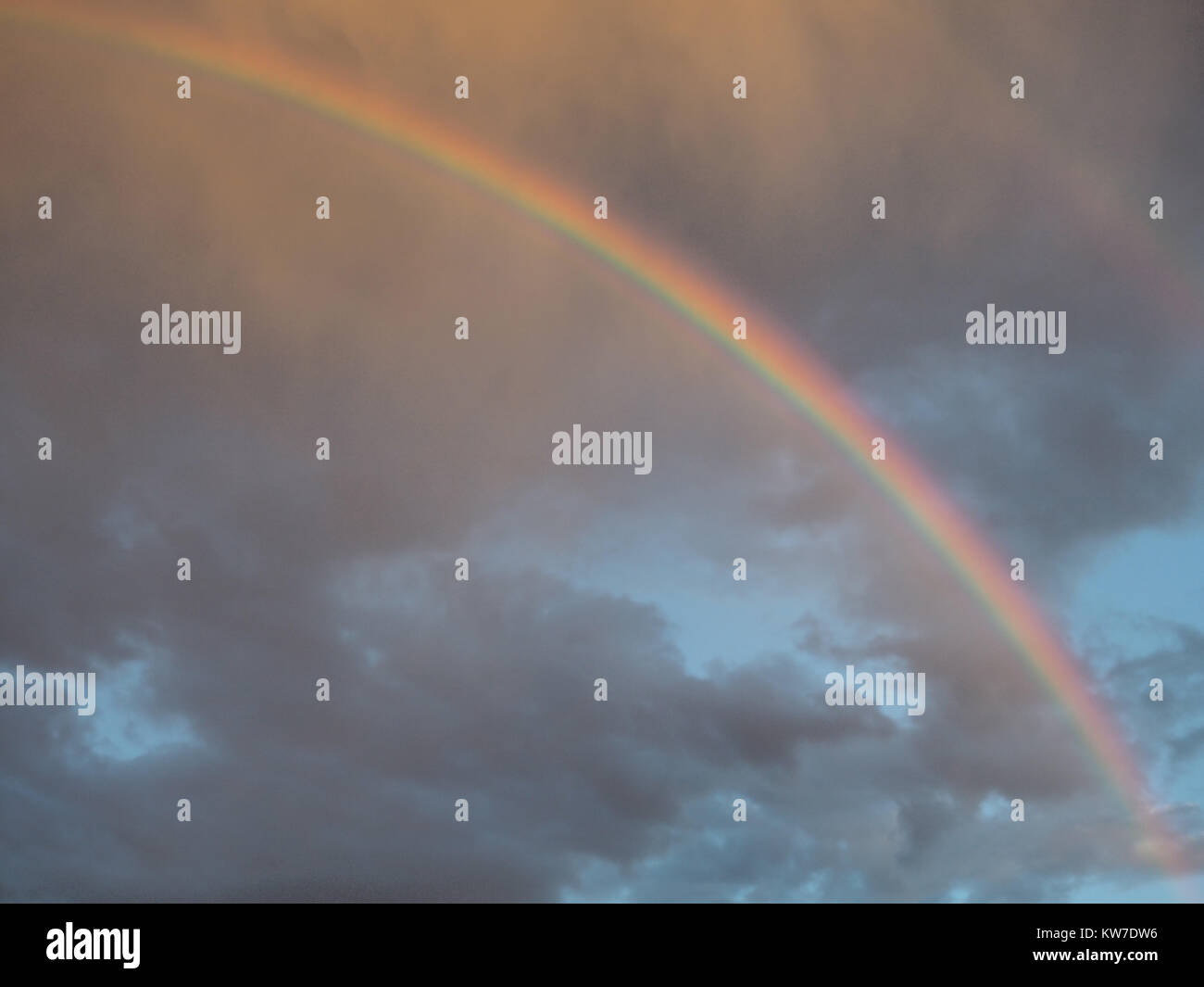 Large colored rainbow in a blue sky amidst gray clouds. Stock Photo