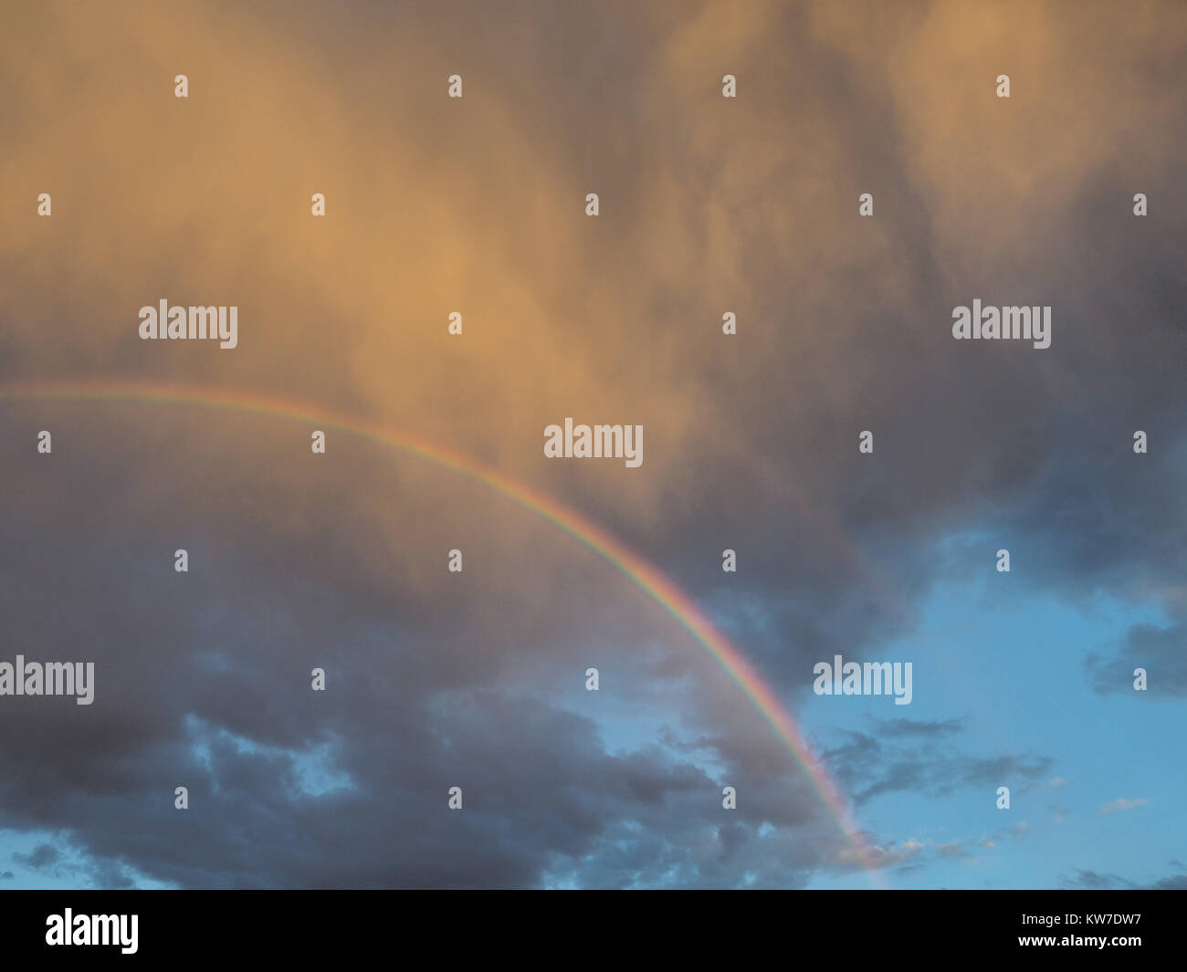 Semicircle of different colors a bright rainbow in a gray cloudy sky, illuminated by the rays of the setting sun. Stock Photo