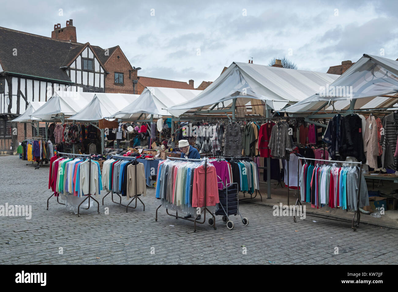 Old lady in historic market place, looks at jumpers hanging on rails by stalls selling clothes - Shambles Market York, North Yorkshire, England, UK. Stock Photo