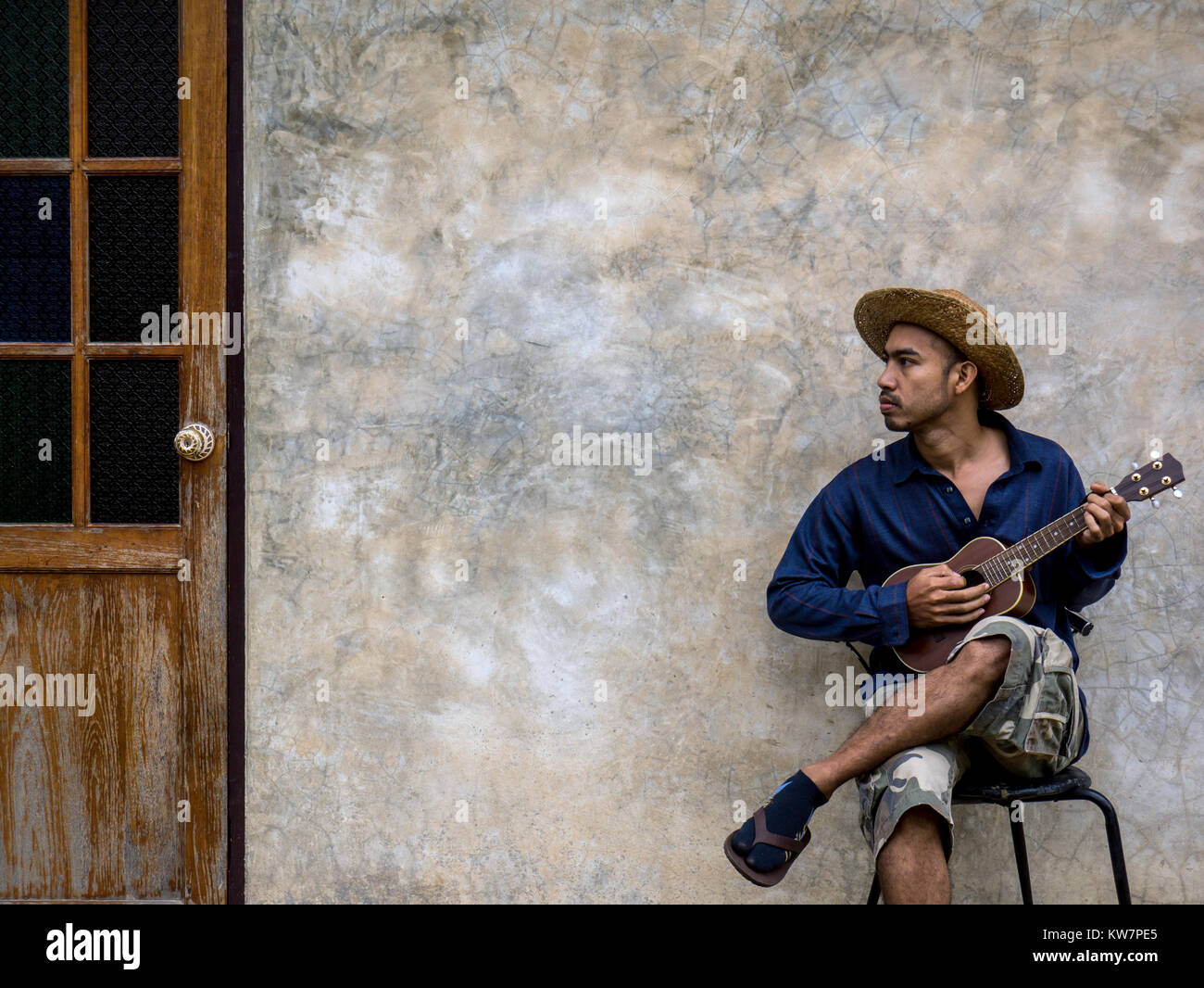 Asian musician man in casual dress sitting on chair and playing ukulele with classic cement wall and wooden door background Stock Photo