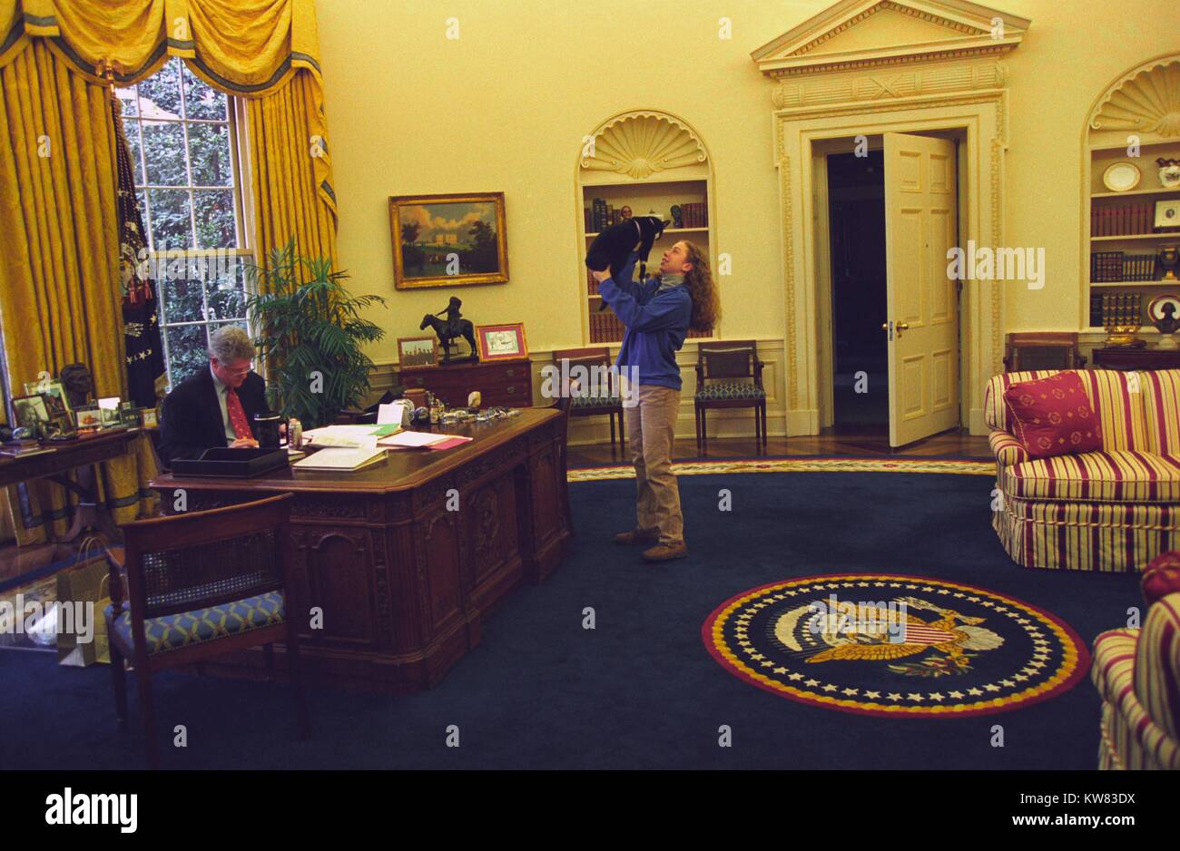 President Bill Clinton, wearing suit and glasses, works at his desk in the Oval Office, while the First Daughter Chelsea Clinton, wearing khaki trousers and a blue sweater, stands facing him with arms stretched upward cradling the First Pet, Socks the Cat, with black fur, white face, and tiny collar, inside the White House, Washington, District of Columbia, December 24, 1994. Stock Photo