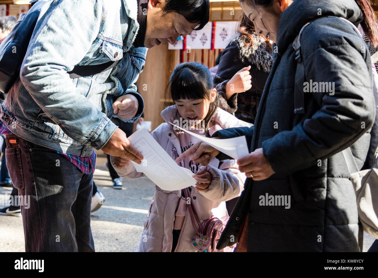 Japan, Nishinomiya Shinto Shrine. New year's day, Shogatsu. Family on Hatsumode visit, (first visit of the new year), reading a fortune paper, Omikuji to their child daughter. She looks happy and smiling as they bend to explain. Stock Photo