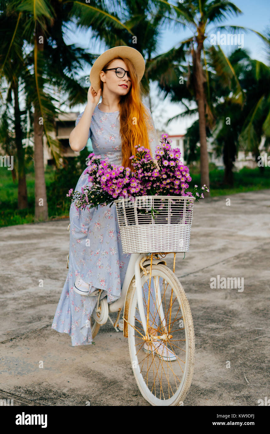Summer vacation. Orange hair woman in dress holding hat, sitting on a bicycle Stock Photo