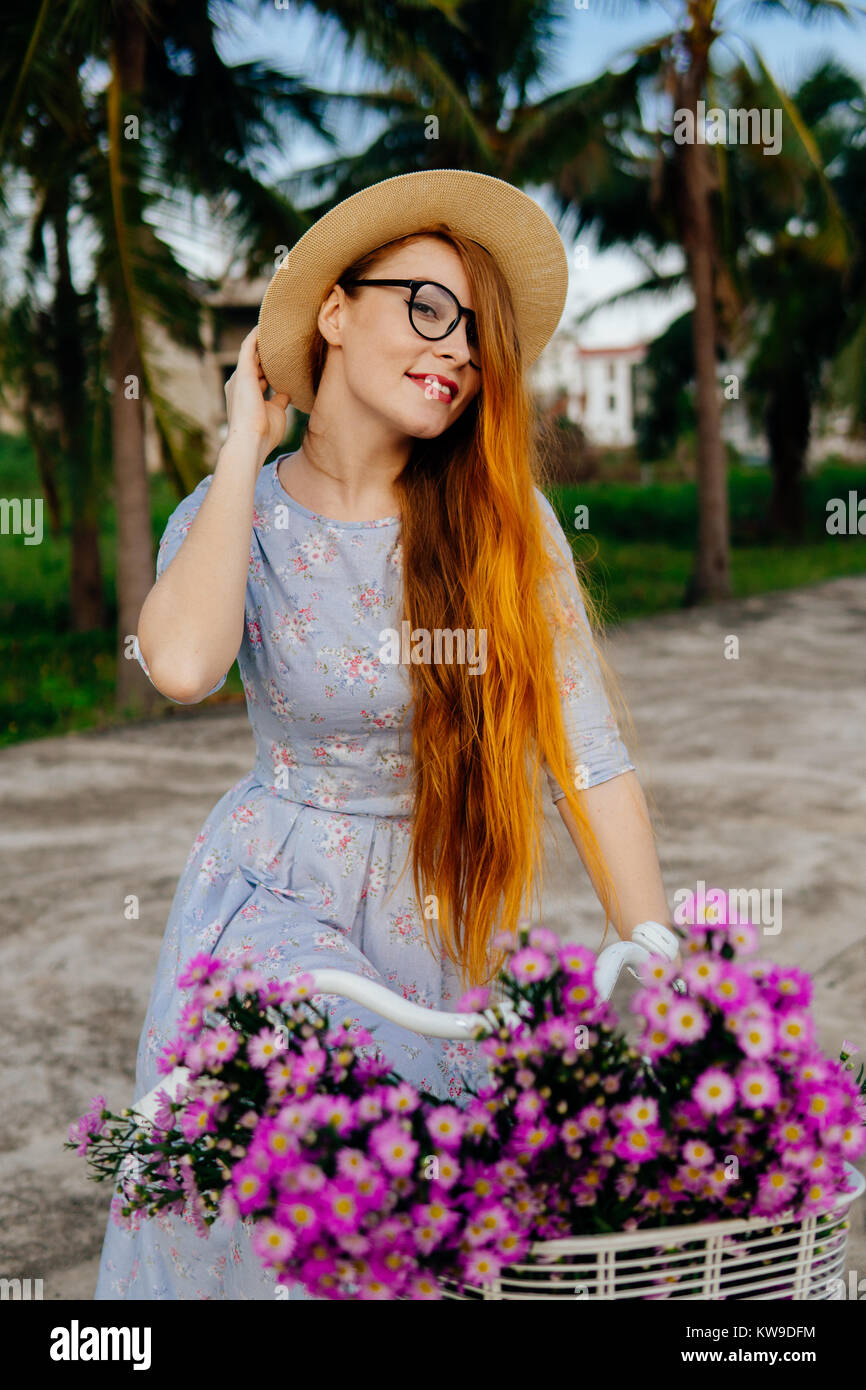 Summer vacation. Orange hair woman in dress holding hat, sitting on a bicycle Stock Photo