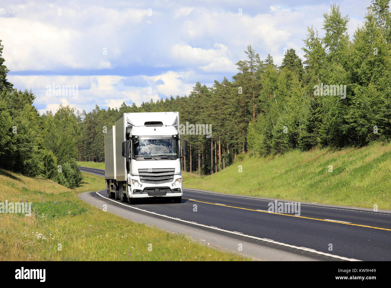 White cargo truck transports goods along highway through green rural scenery on a beautiful day of summer. Stock Photo