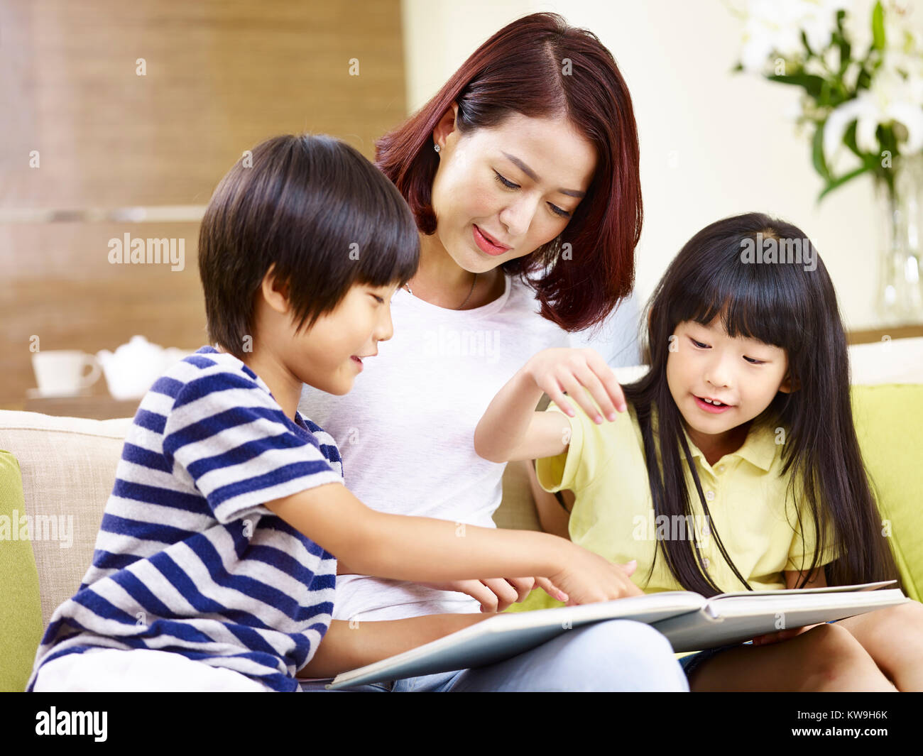 young asian mother and her son and daughter sitting on couch reading a book together. Stock Photo