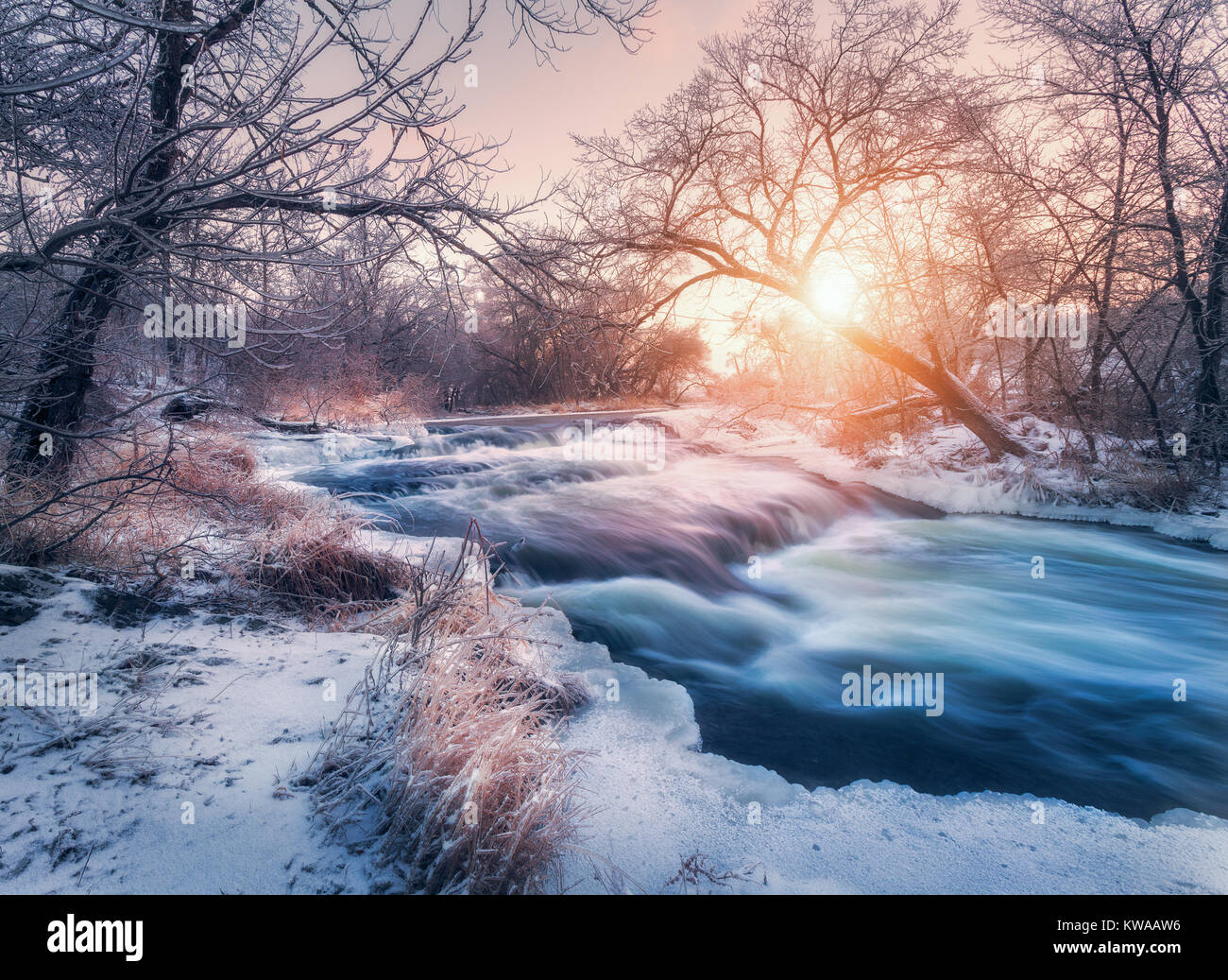 Winter forest with amazing river at sunset. Winter landscape with snowy trees, ice, beautiful frozen river, snowy bushes, colorful sky in dusk. Blurre Stock Photo