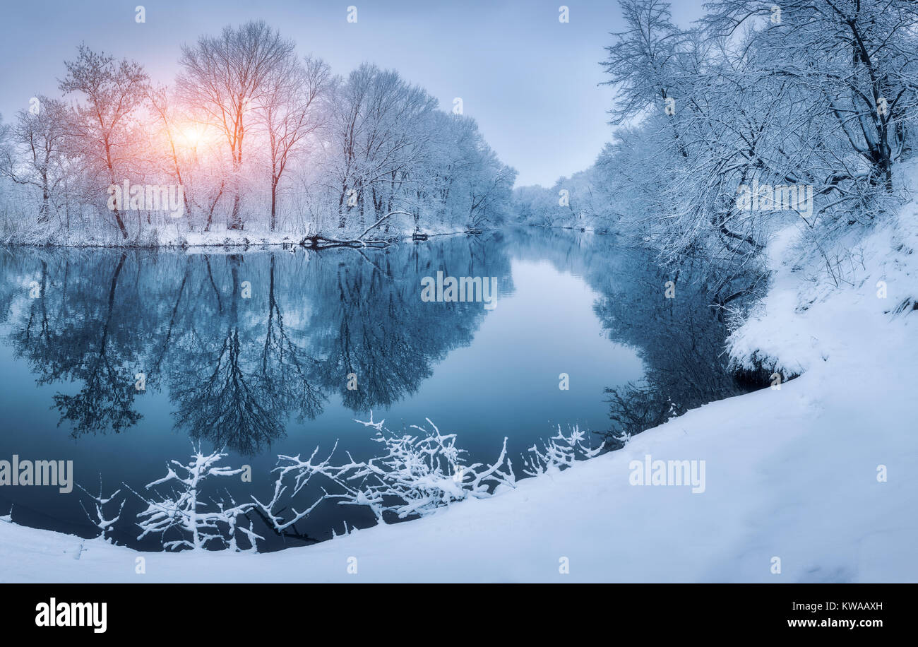 Winter forest on the river at sunset. Colorful landscape with snowy trees, river with reflection in water in cold evening. Snow covered trees, lake, s Stock Photo