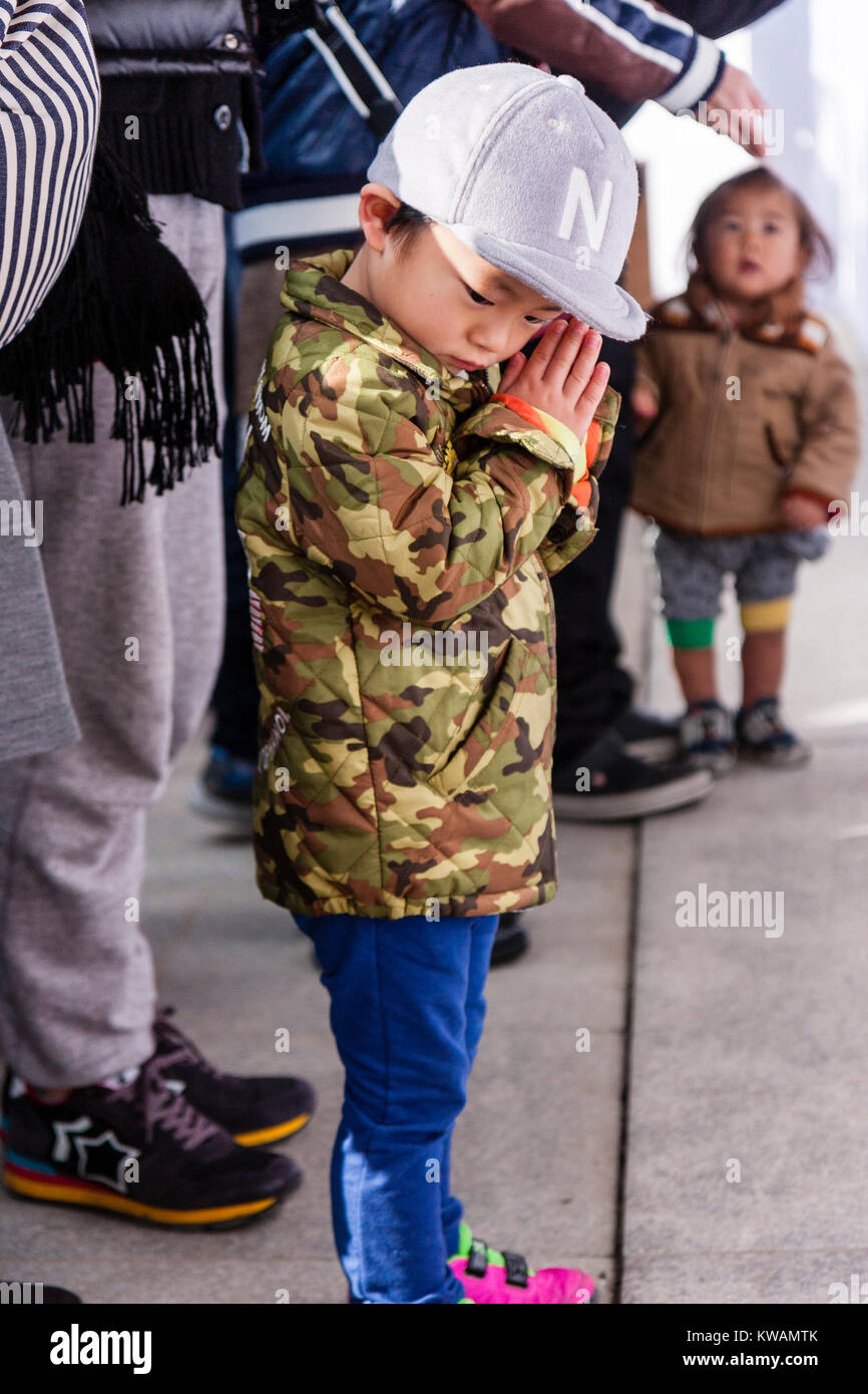 Japan, Nishinomiya Shinto shrine. Hatsumode, first visit of the new year. Little boy, child, about five to six years old, standing in front of Haiden praying but glancing sideways. Side view. Daytime. Wears baseball cap. Stock Photo