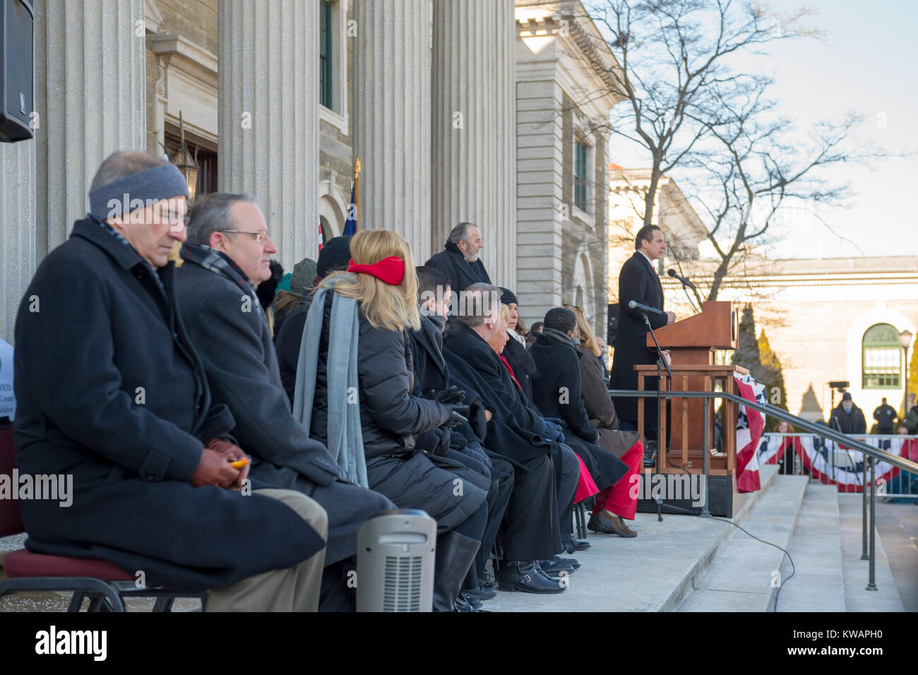 Mineola, United States. 01st Jan, 2018. Mineola, New York, USA. January 1, 2018. New York Governor ANDREW CUOMO is speaking at poidum during historic swearing-In of Laura Curran as Nassau County Executive, the first female County Executive. Cuomo administered the Oath of Office to Curran. Nassau County Legislators are seated left of Cuomo. Temperature was a freezing 14 ℉ Fahrenheit/-10 ℃ Celsius for the outdoor ceremony held in front of Theodore Roosevelt Executive & Legislative Building. Standing behind Cuomo is Mayor FRANCIS MURRAY of Rockville Centre. Credit: Ann E Parry/Alamy Live News Stock Photo