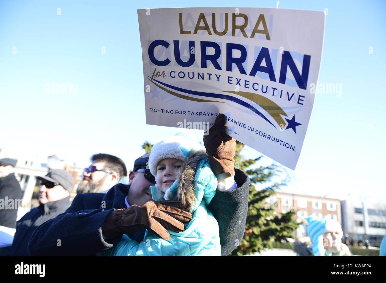 Mineola, New York, USA. January 1, 2018. JOE CAVANAUGH, of Oceanside, is holding his young daughter EMMA CAVANAGH and a political poster 'Laura Curran for County Executive' while standing bundled up in warm winter coats in audience during historic swearing-In of Laura Curren as Nassau County Executive, the first female County Executive, held outdoors. Temperature was a freezing 14 ℉ Fahrenheit / -10 ℃  Celsius for the outdoor ceremony held in front of Theodore Roosevelt Executive & Legislative Building. Stock Photo