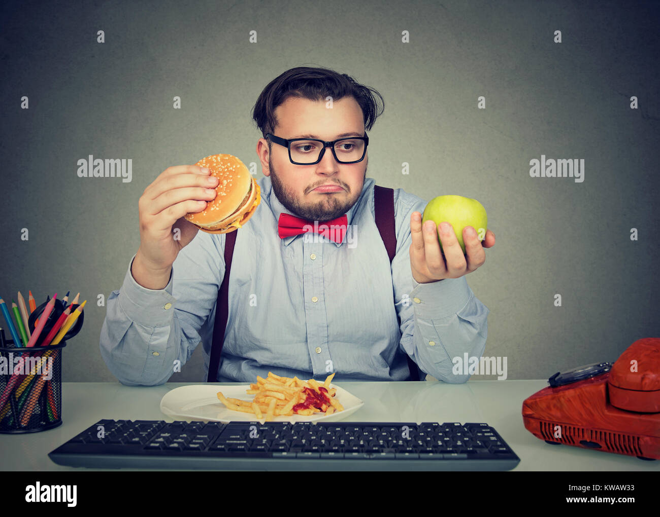 Chubby man looking confused and sad trying to choose between apple and burger while spending time in office. Stock Photo