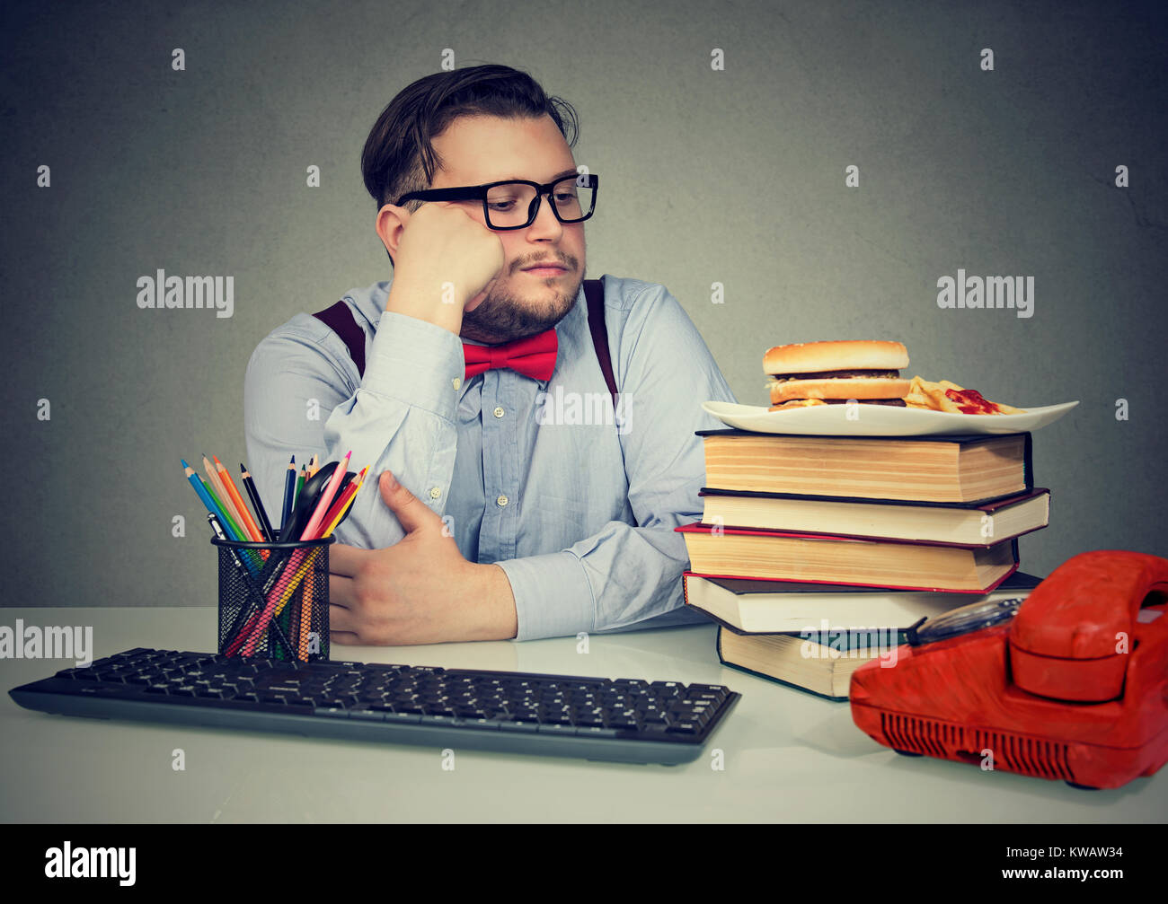 Chunky man sitting at working table and looking at burger with temptation having stress. Stock Photo
