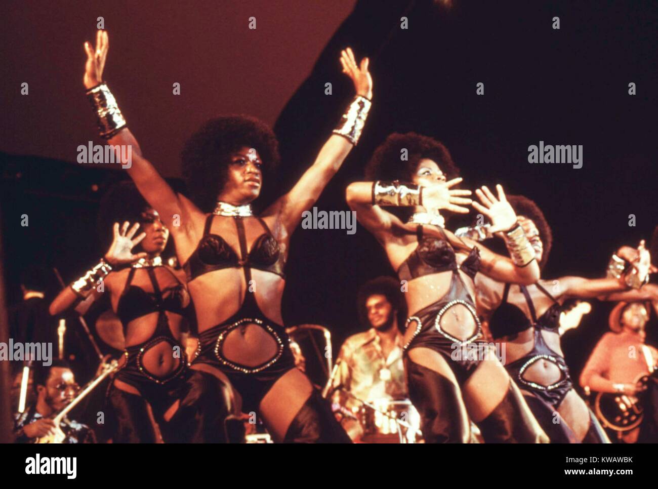 During an annual Black Talent Expo, female dancers for soul singer Isaac Hayes shine on stage in black leather body suits, tall boots, shiny cuffs, and Afros, stretching out their hands toward the ceiling, or pulling them inward with palms facing out, a boisterous band playing behind them under the room's bright spotlight, Chicago, Illinois, 1973. Image courtesy National Archives. Stock Photo