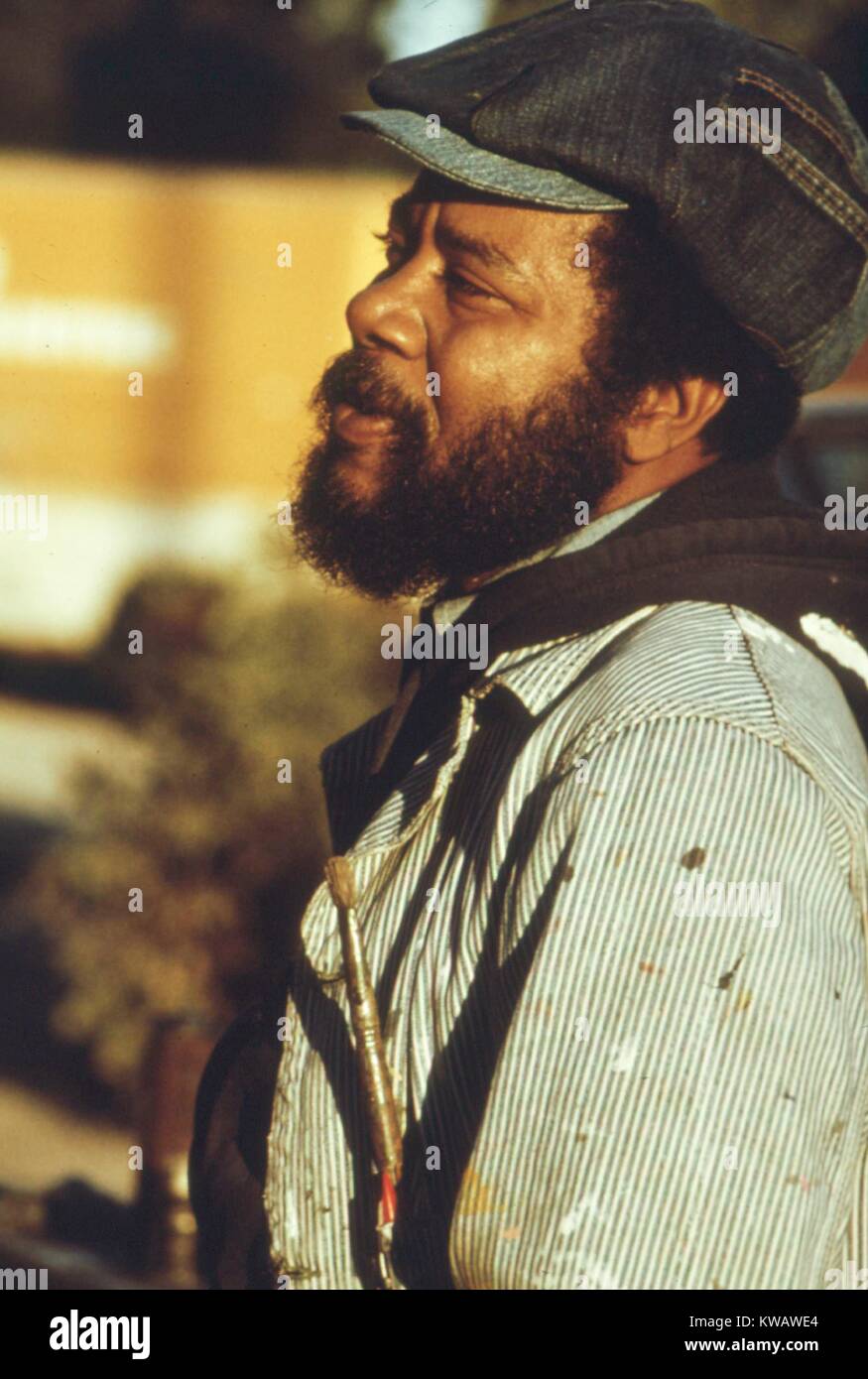 Outside at 33rd and Giles Street in the South Side, a muralist with a beard, newsboy hat, and denim jacket speckled with paint stands in profile gazing toward his day's worth of work as the warm sun sets, Chicago, Illinois, 1973. Image courtesy National Archives. Stock Photo
