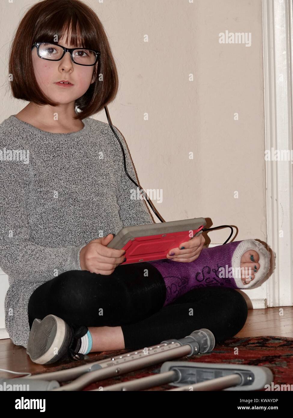 Young girl wearing glasses, with cast on broken foot, sitting cross legged on floor holding electronic tablet. Facing camera with crutches at feet. Stock Photo