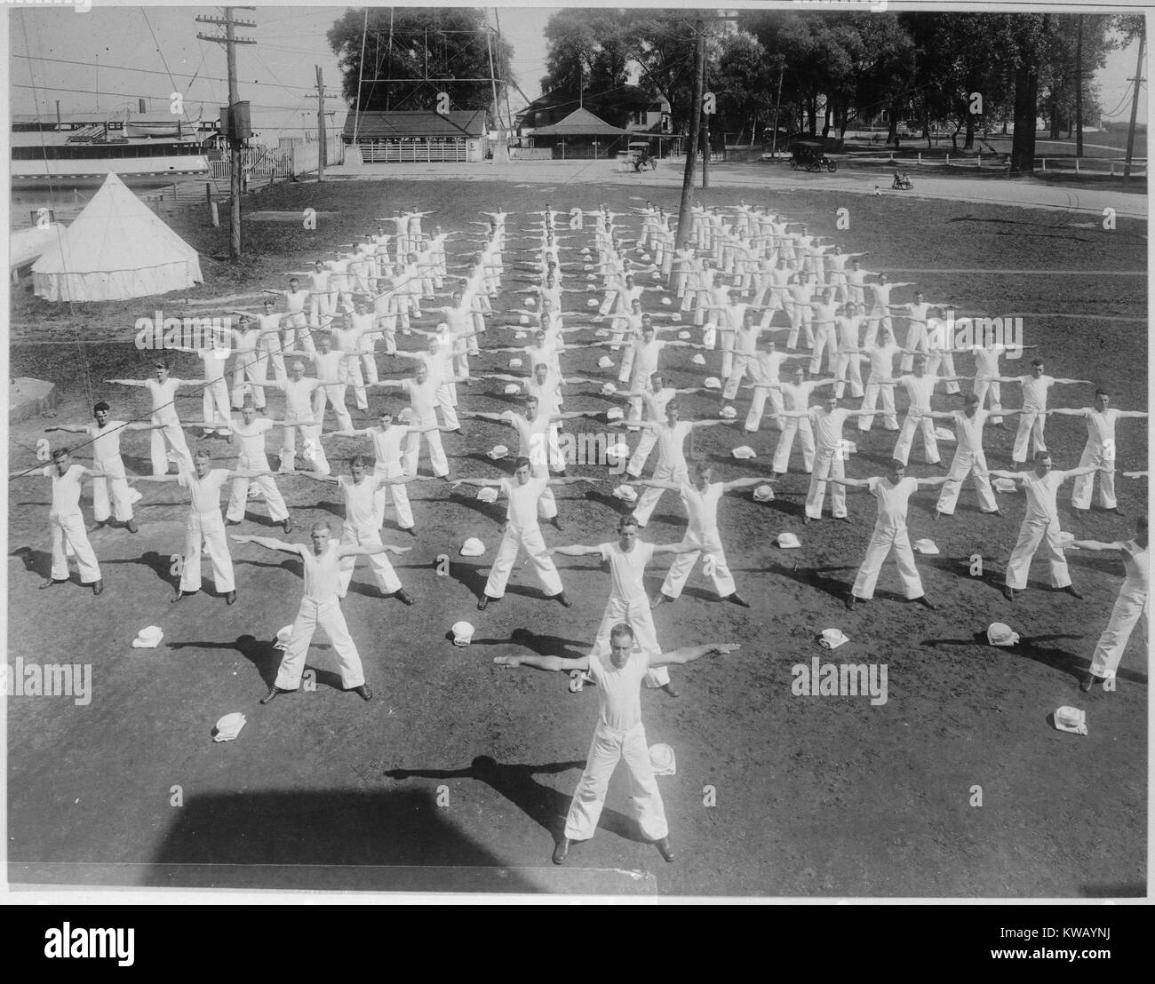 Dozens of male recruits at a Naval Militia Camp stand in formation wearing white tee shirts and pants, arms outstretched doing jumping jacks during their outdoor exercise routine, Somersville, New York, 1917. Image courtesy National Archives. Stock Photo