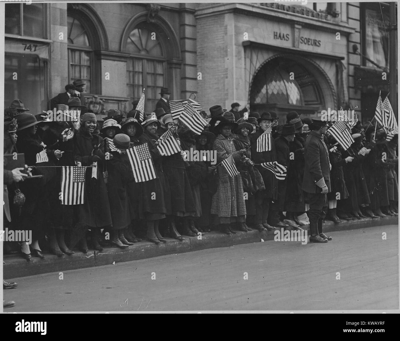 Smiling and waving United States flags while crowded on the sidewalk of the street, onlookers dressed in coats and hats await the parade of the famous 369th African American Infantry, formerly the 15th New York regulars, New York, New York, 1917. Image courtesy National Archives. Stock Photo
