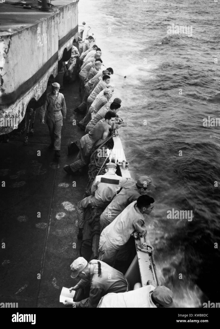 U.S. Marines stand along the rail of the USS Clymer taking them to Korea in late July, 1950. To the aft a Marine is washing his dungarees by dragging them along behind the ship. Korean War, 1950-53. (BSLOC 2014 11 11) Stock Photo