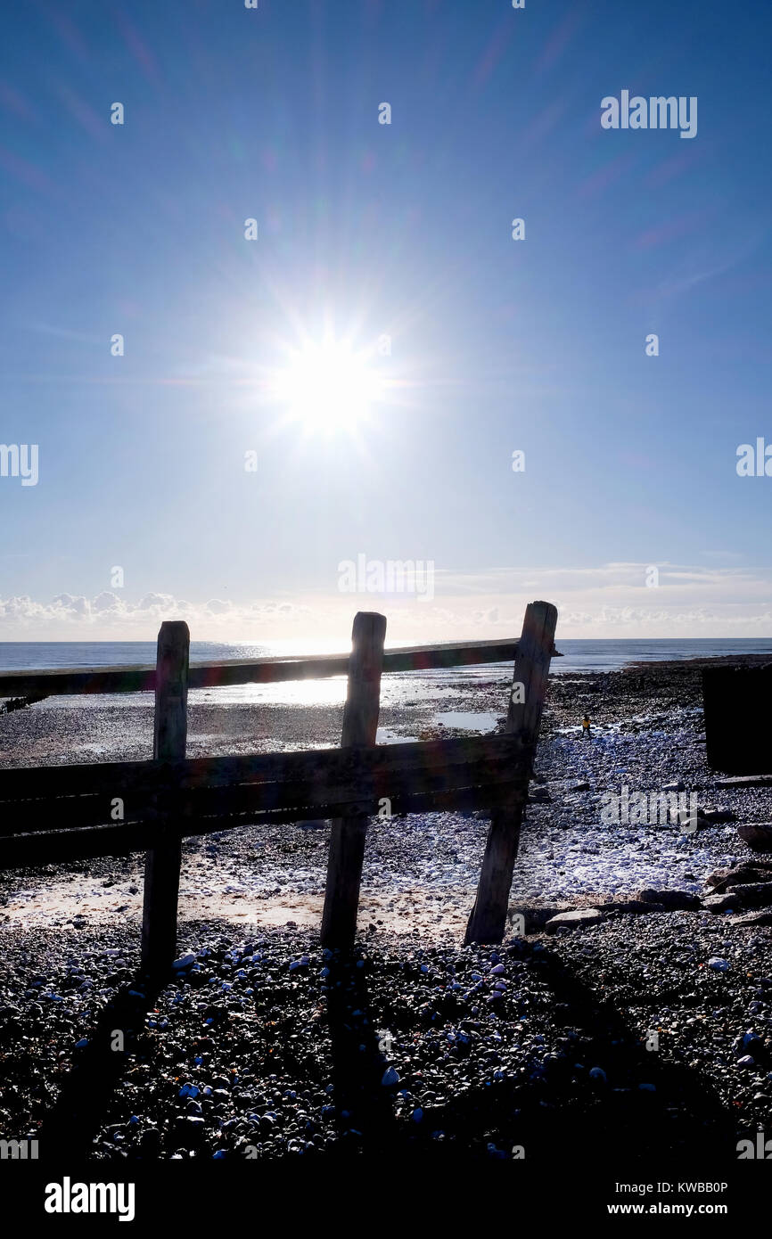 beach with rocks and pebbles and an old wooden big sea defence fence, the sea is behing and the sun is low in the sky and shining directly at the came Stock Photo