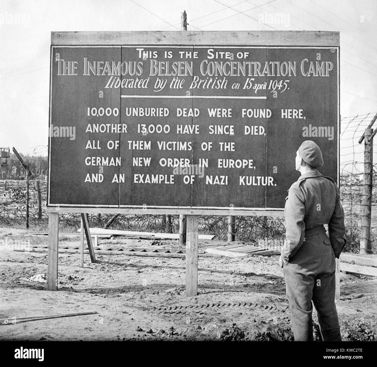 British soldier stands by a sign posted at the site of the Belsen Concentration Camp, 1945. Sign reads: 10,000 unburied dead were found here, another 13,000 have since died, all of them victims of the German new order in Europe, and an example of Nazi Kultur. World War 2 (BSLOC 2015 13 11) Stock Photo