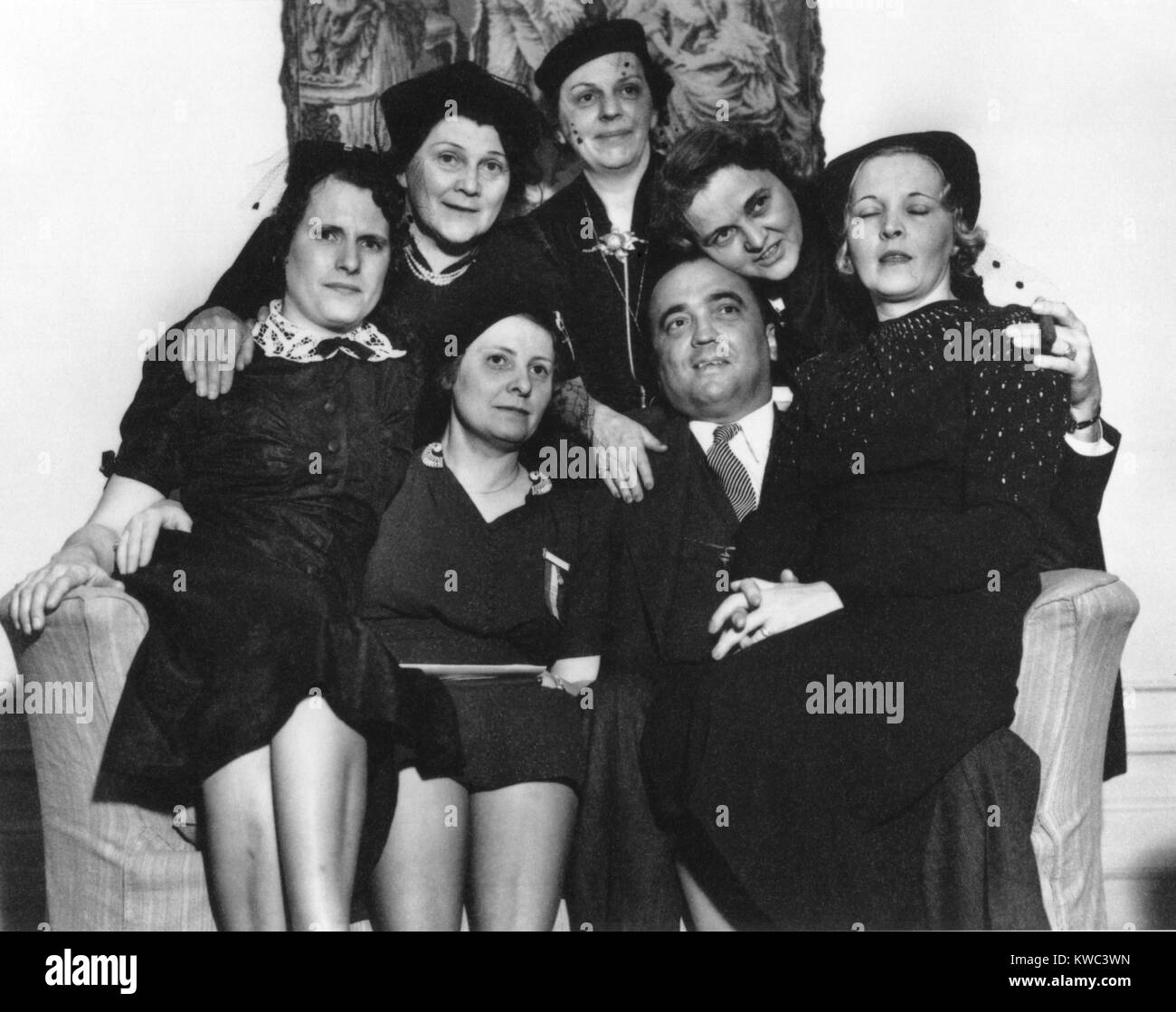 FBI Director J. Edgar Hoover on a loveseat with six women at the Willard Hotel, Washington, D.C. They were attending the convention of the International Association for Identification, for professionals in forensic identification, investigation, and scientific examination of physical evidence. Ca. 1940. (BSLOC_2015_14_20) Stock Photo