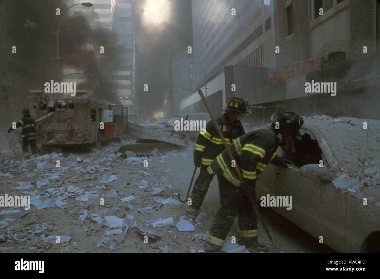 NYC firemen checking a car on Barclay Street after the 9-11 terrorist attack on World Trade Center. In the mid-ground fireman pulls a water hose from fire truck. View is toward West Broadway and includes WTC 7, on right, clad in red exterior masonry. WTC 7 collapsed at 5:20 PM on Sept. 11, 2001, due to fires ignited by debris from the North Tower (WTC 1) destruction earlier in the day. (BSLOC 2015 2 53) Stock Photo
