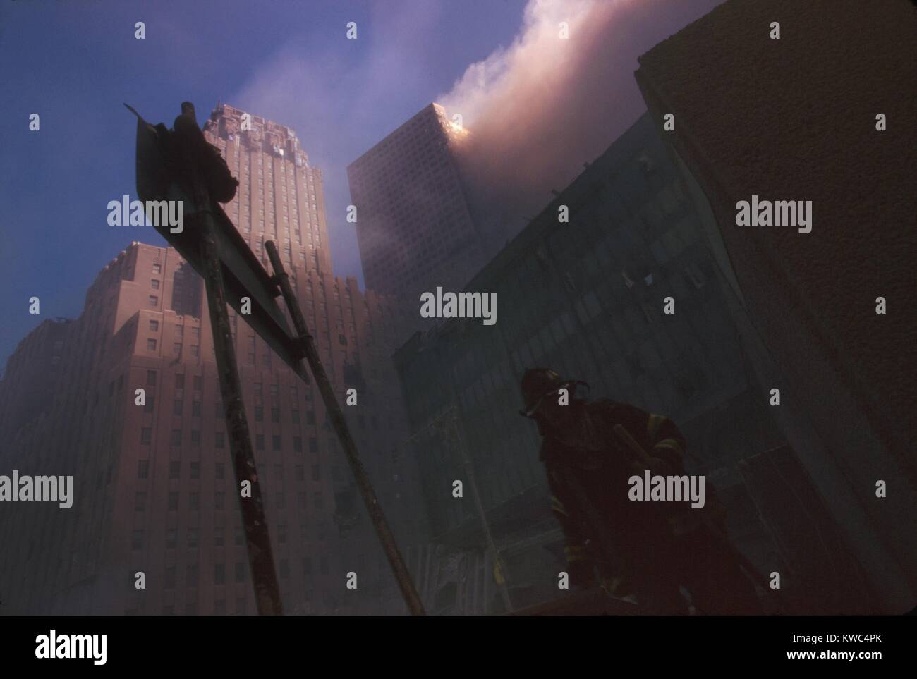 Fire fighter at base of burning WTC 6 following the North Tower (WTC 1) collapse. Dense dust and smoke block sunlight. At far left is the New York Telephone Company Building, built in the 1920s, damaged but repairable. In upper center is the damaged and burning WTC 7, which will collapse at 5:20 on Sept. 11, 2001. (BSLOC 2015 2 59) Stock Photo