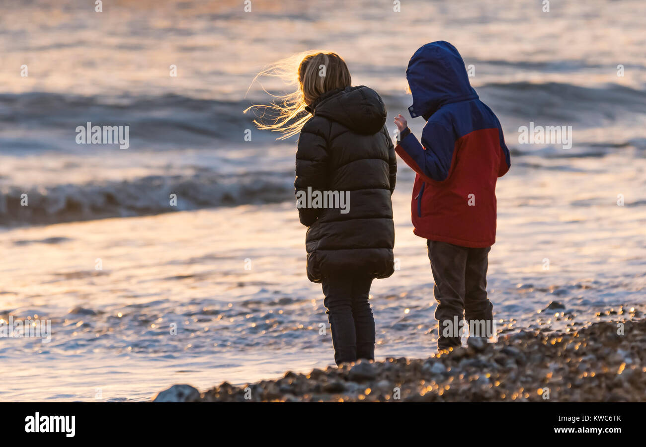 Young male and female children standing by the sea wearing coats on a Winter evening in the UK. Stock Photo