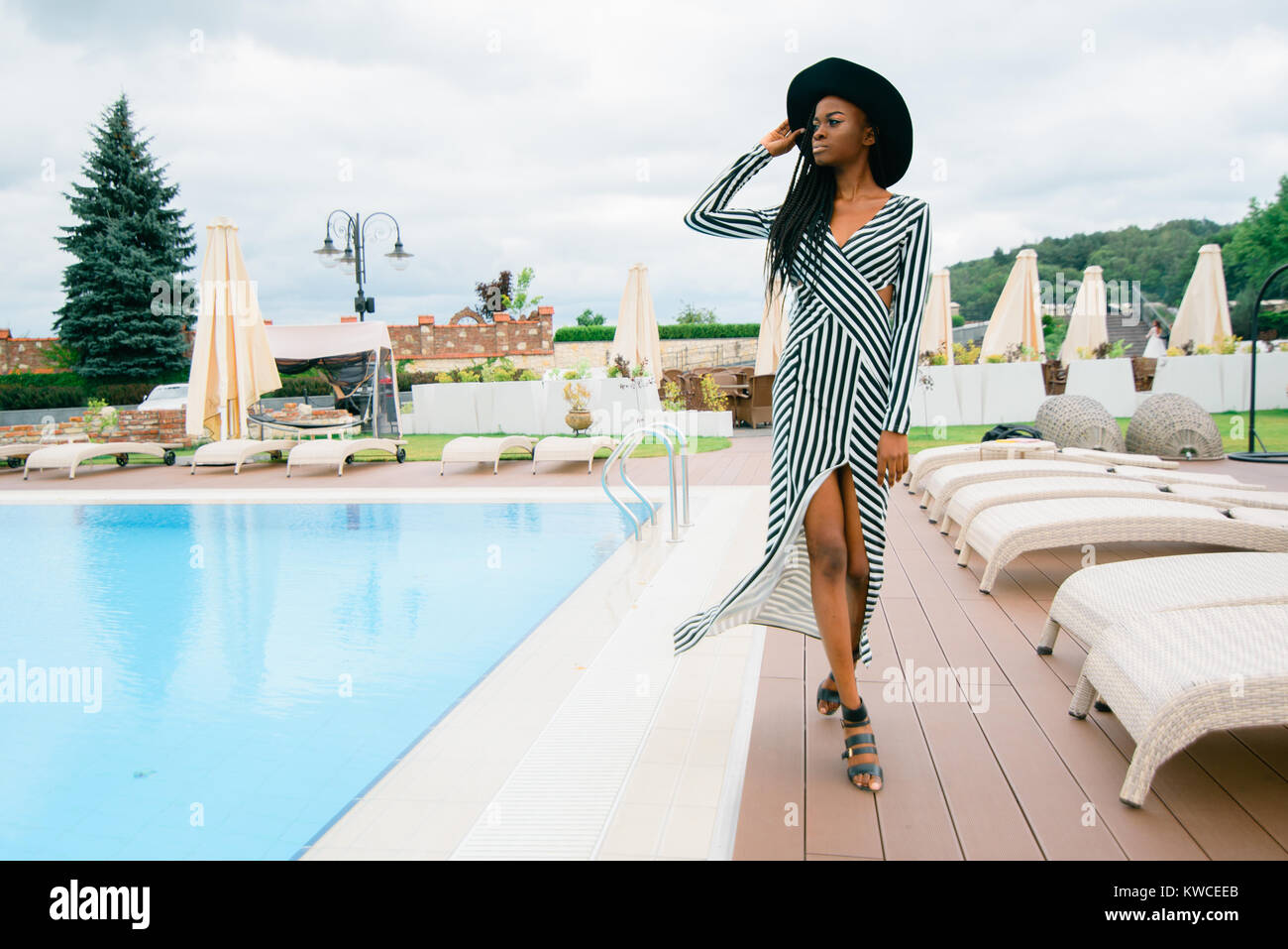 Attractive, pretty, tender, beautiful american woman in long dress in stripes and black hat. Model walking near pool of luxury hotel in Asia. Stock Photo
