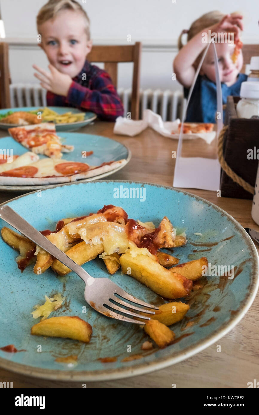 A half eaten plate of cheesy chips with ketchup on a table in front of children eating food. Stock Photo